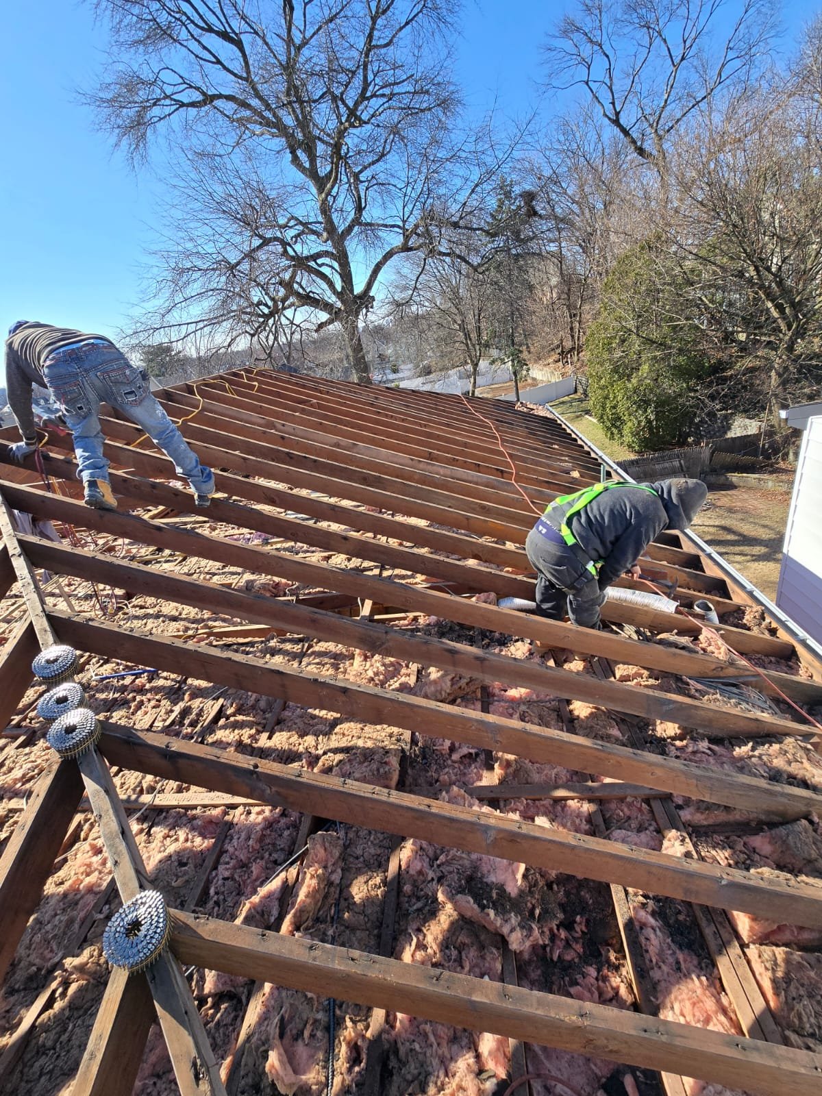 Two construction workers installing roofing framework on a house, with trees and a clear blue sky in the background.
