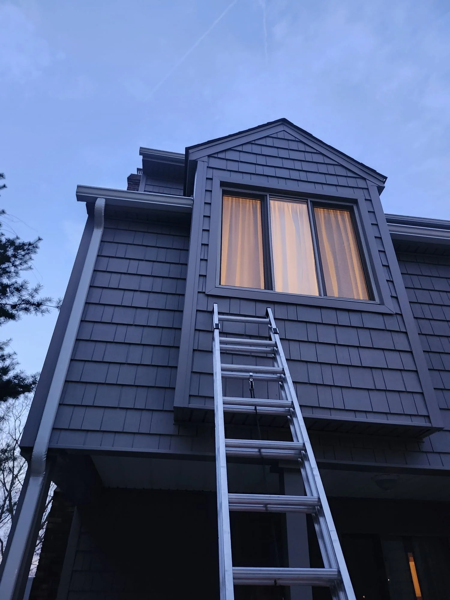 A house with gray siding and a large window with curtains, a metal ladder leaning against it, and a clear sky with some clouds and a contrail.