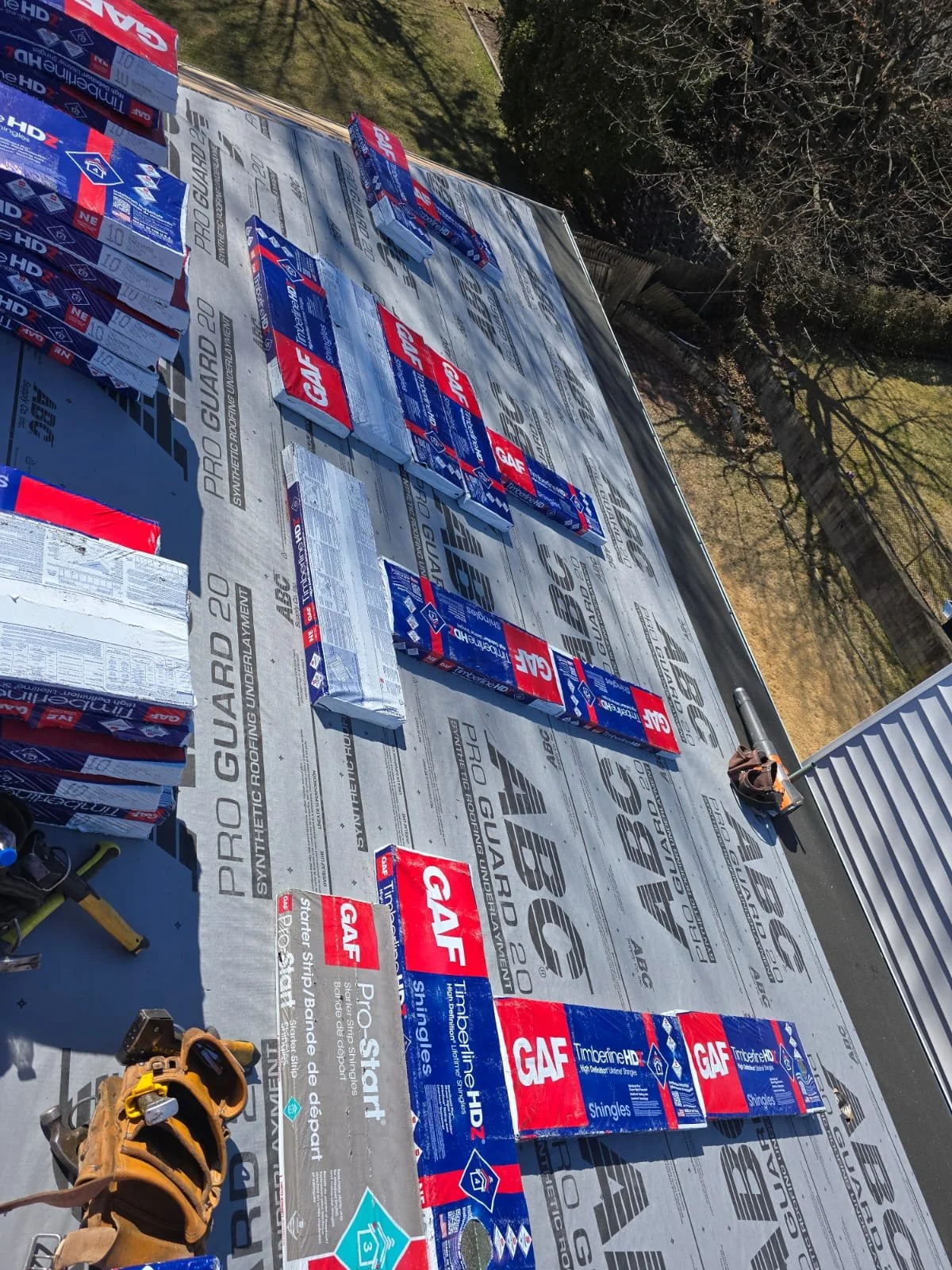 Roof underlayment with numerous GAF Timberline HD shingles packages and tools laid out for roofing installation. The background shows a yard with trees and a wooden fence.