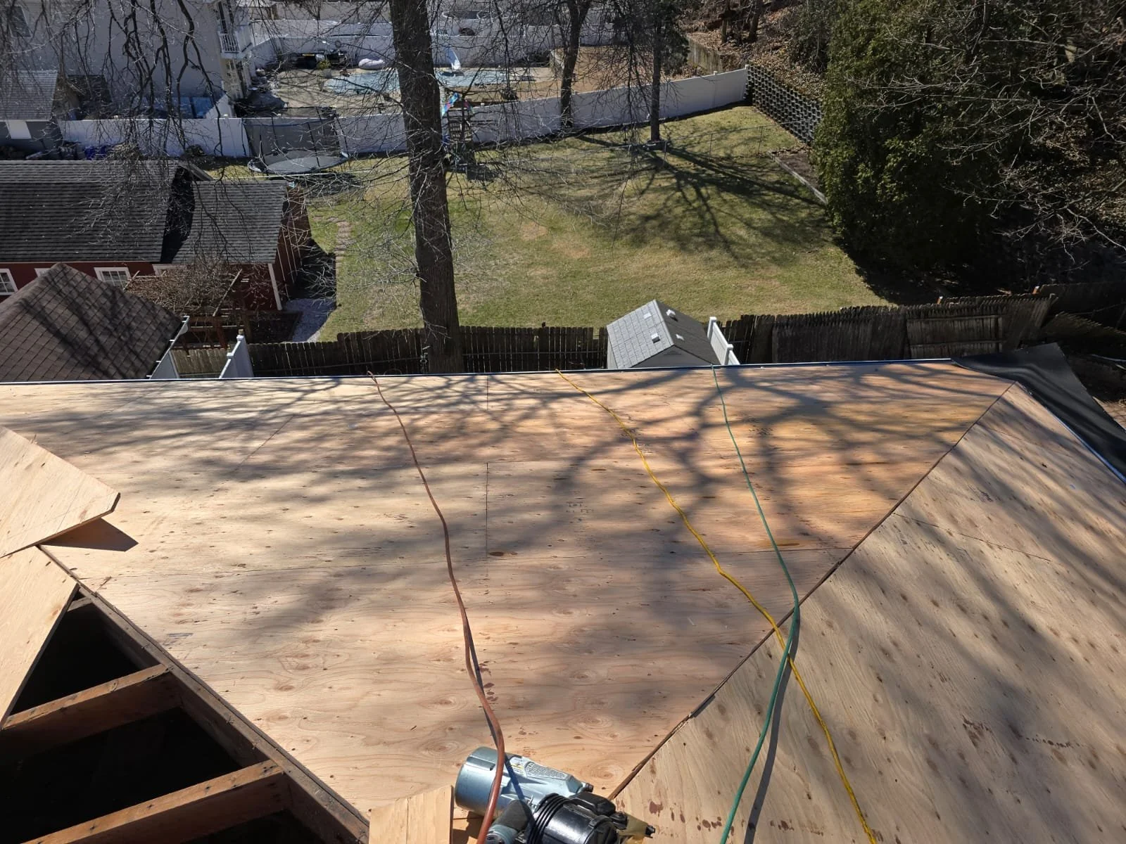 View from a rooftop under construction, showing unfinished wooden surface with electrical wires, over a backyard with grass, trees, and neighboring houses.