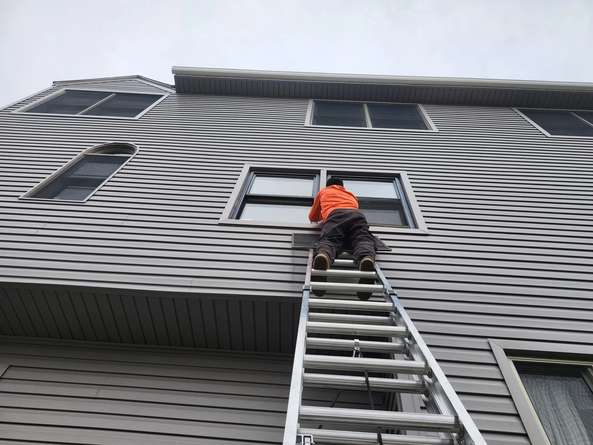 A person wearing an orange jacket and black pants is on a ladder, cleaning or working on the exterior window of a multi-story building with vinyl siding.