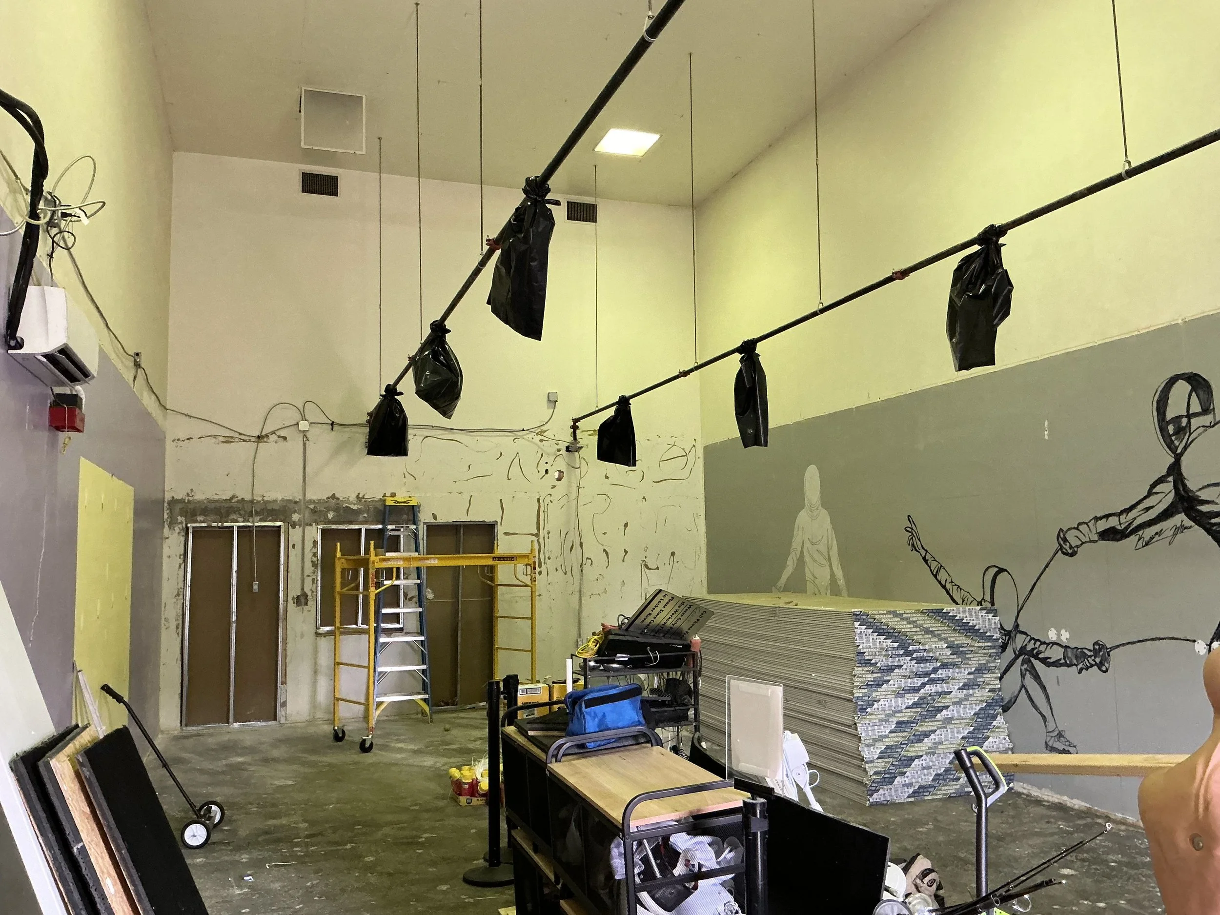 Construction or art studio with yellow walls, scaffolding, tools, and artwork, including sketches of people on the walls and hanging black bags.