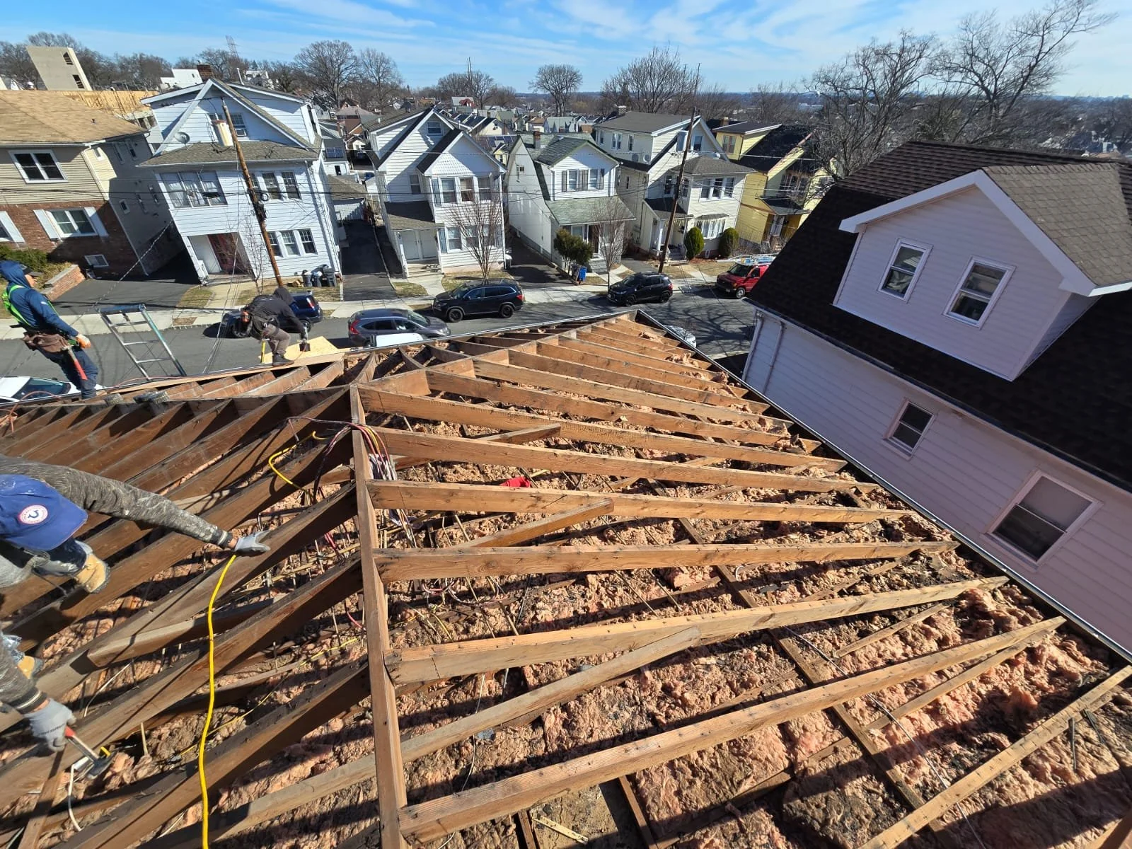 Construction workers installing roof trusses on a house during daytime, with a background of residential neighborhood houses and a partly cloudy sky.