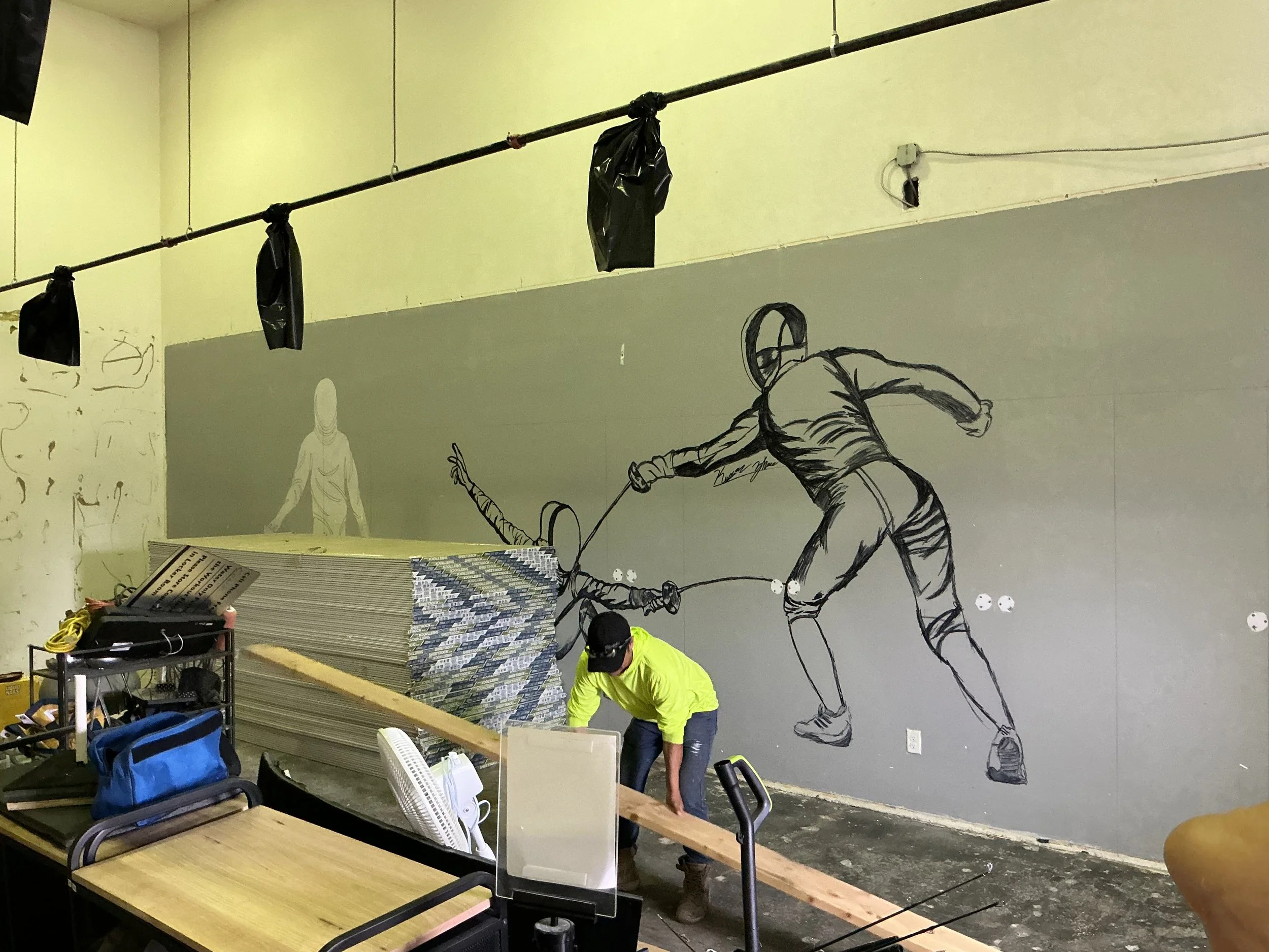 A large wall mural depicts a man with a stick in various action poses, painted in black and white. Construction materials, a person wearing a neon yellow jacket and black cap, and other items are in the foreground, indicating a workspace or studio.