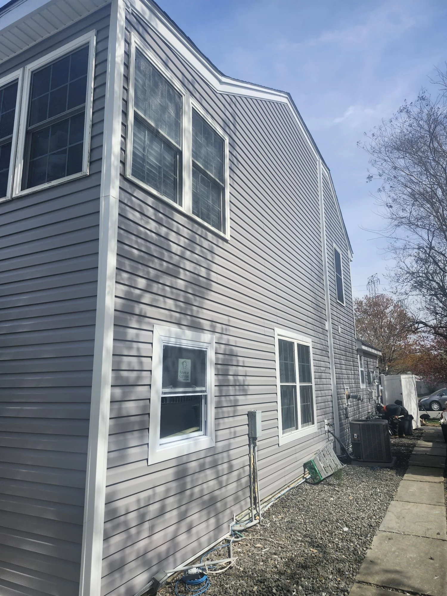 Side view of a house with gray vinyl siding and white-framed windows, with a concrete sidewalk, gravel yard, and HVAC unit outside on a sunny day.