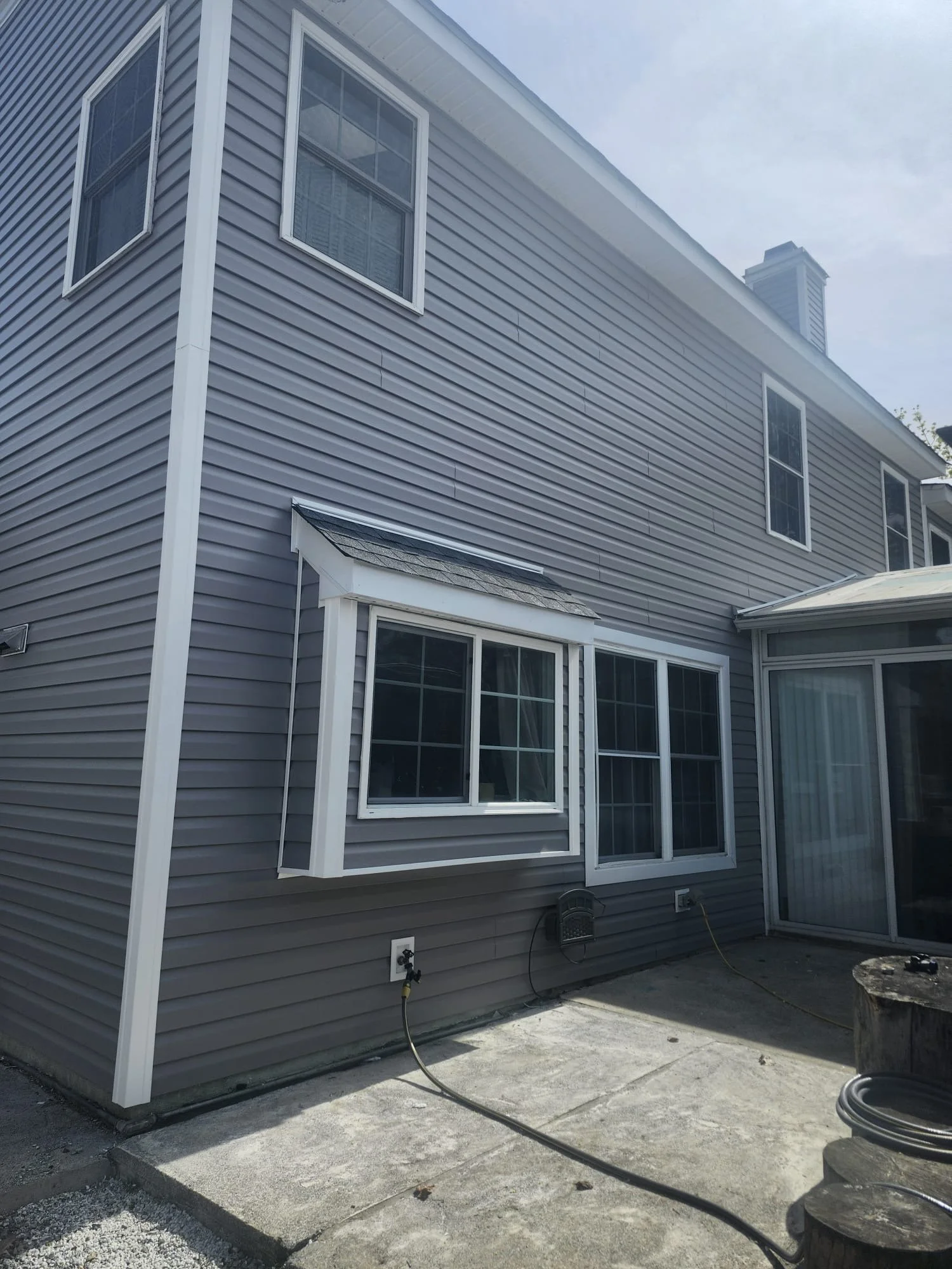 Exterior view of a house with gray vinyl siding, three windows, a small window with a awning, and a concrete patio area with some construction materials and a hose.