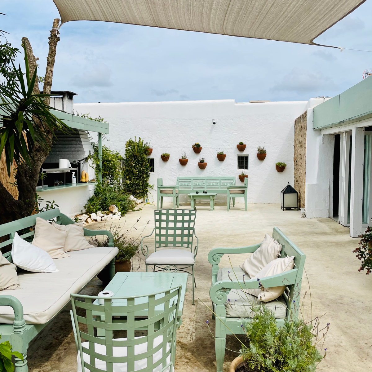 Outdoor patio with mint green furniture, including a bench, chairs, and a table, decorated with pillows and surrounded by potted plants. There are white walls with hanging flower pots and a shade sail overhead, creating a cozy, garden-like setting.
