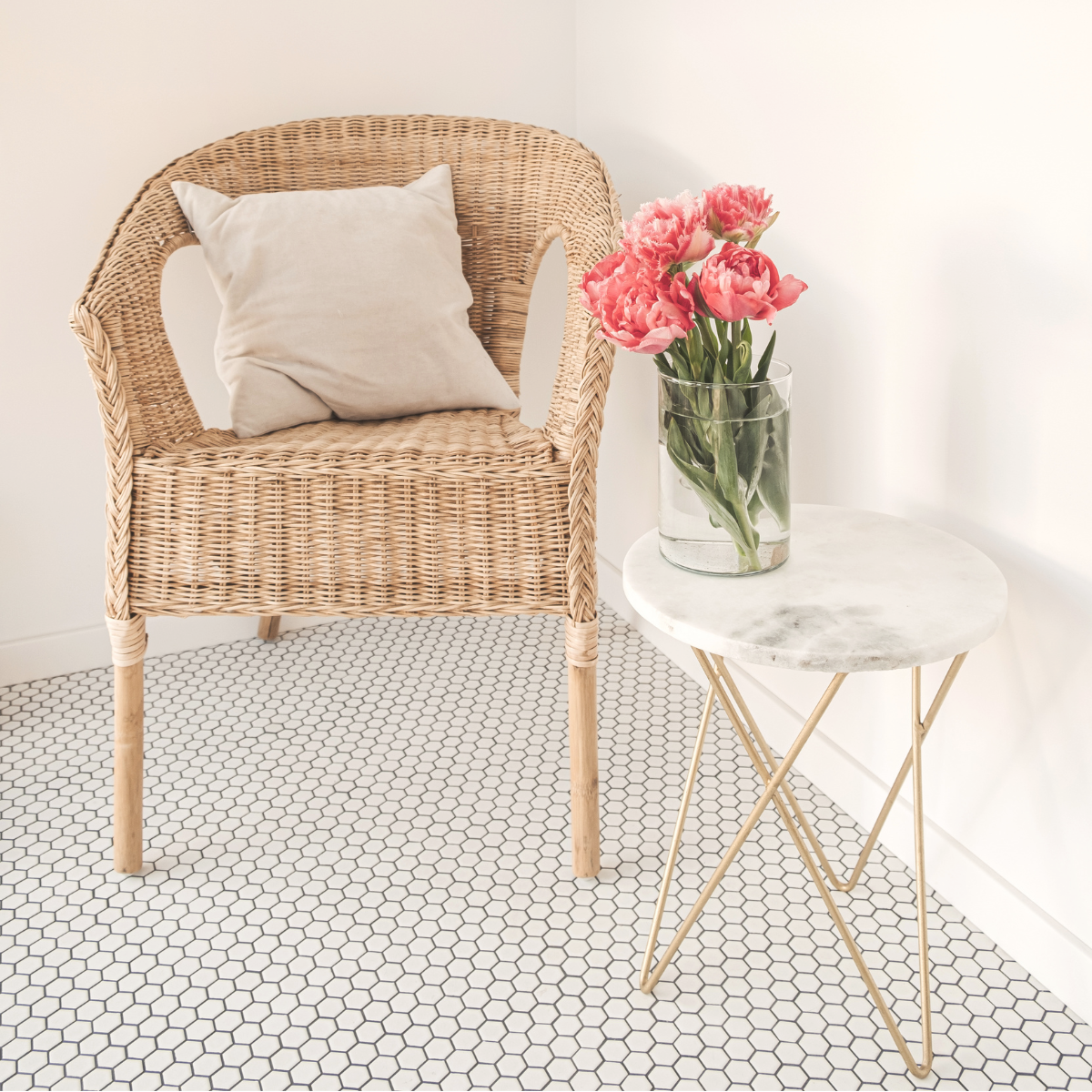 A woven rattan armchair with a beige cushion, next to a round marble-top side table with gold legs, holding a glass vase of pink tulips, in a minimalist room with white walls and black hexagonal tile flooring.