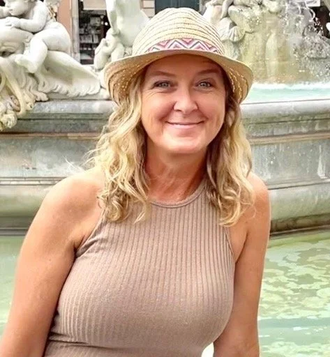 Woman with blonde curly hair wearing a straw hat and beige sleeveless top smiling at the camera in front of a fountain with sculptures.