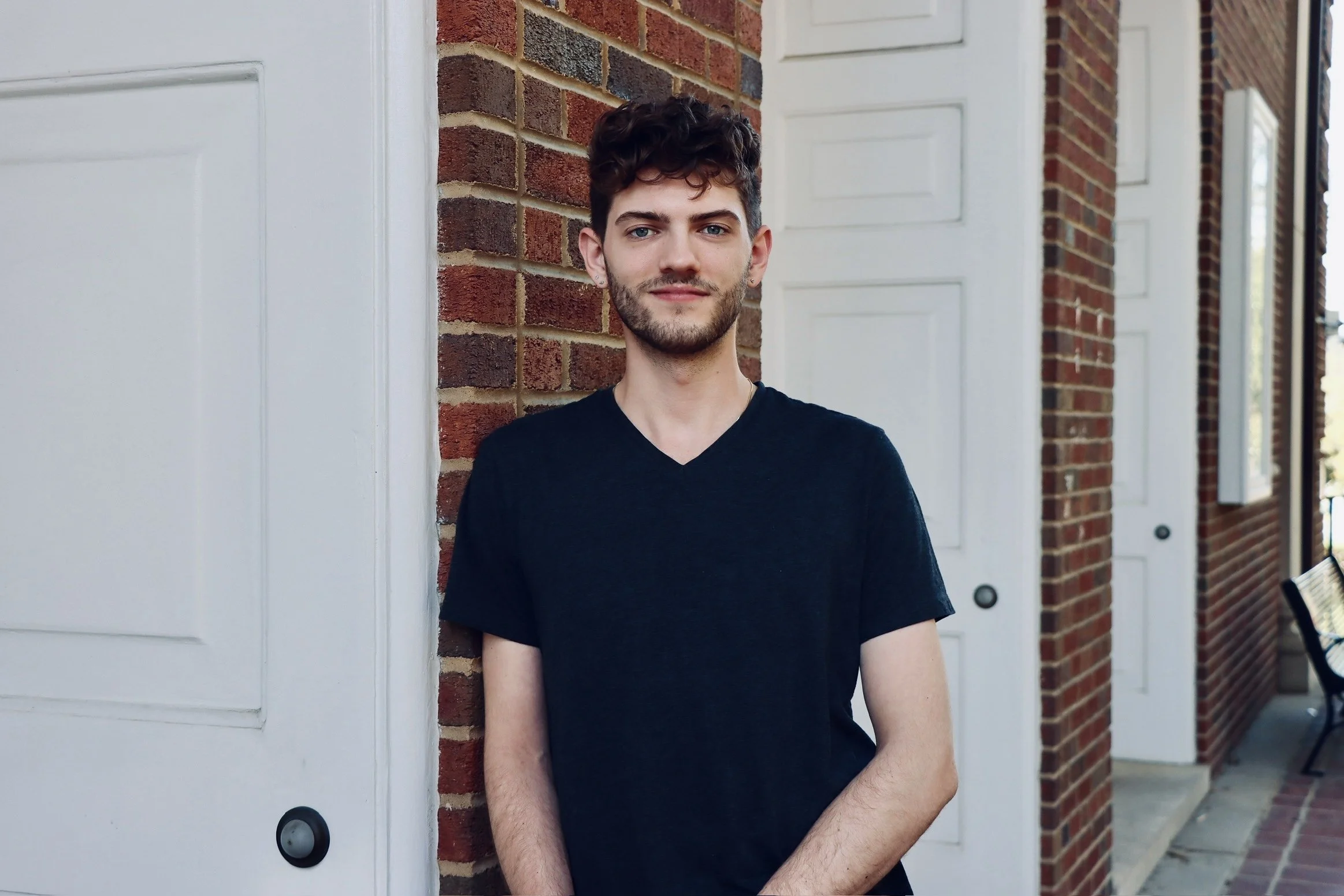 Young man with dark curly hair and a beard, wearing a black V-neck T-shirt, standing outdoors against a brick wall and white doors.