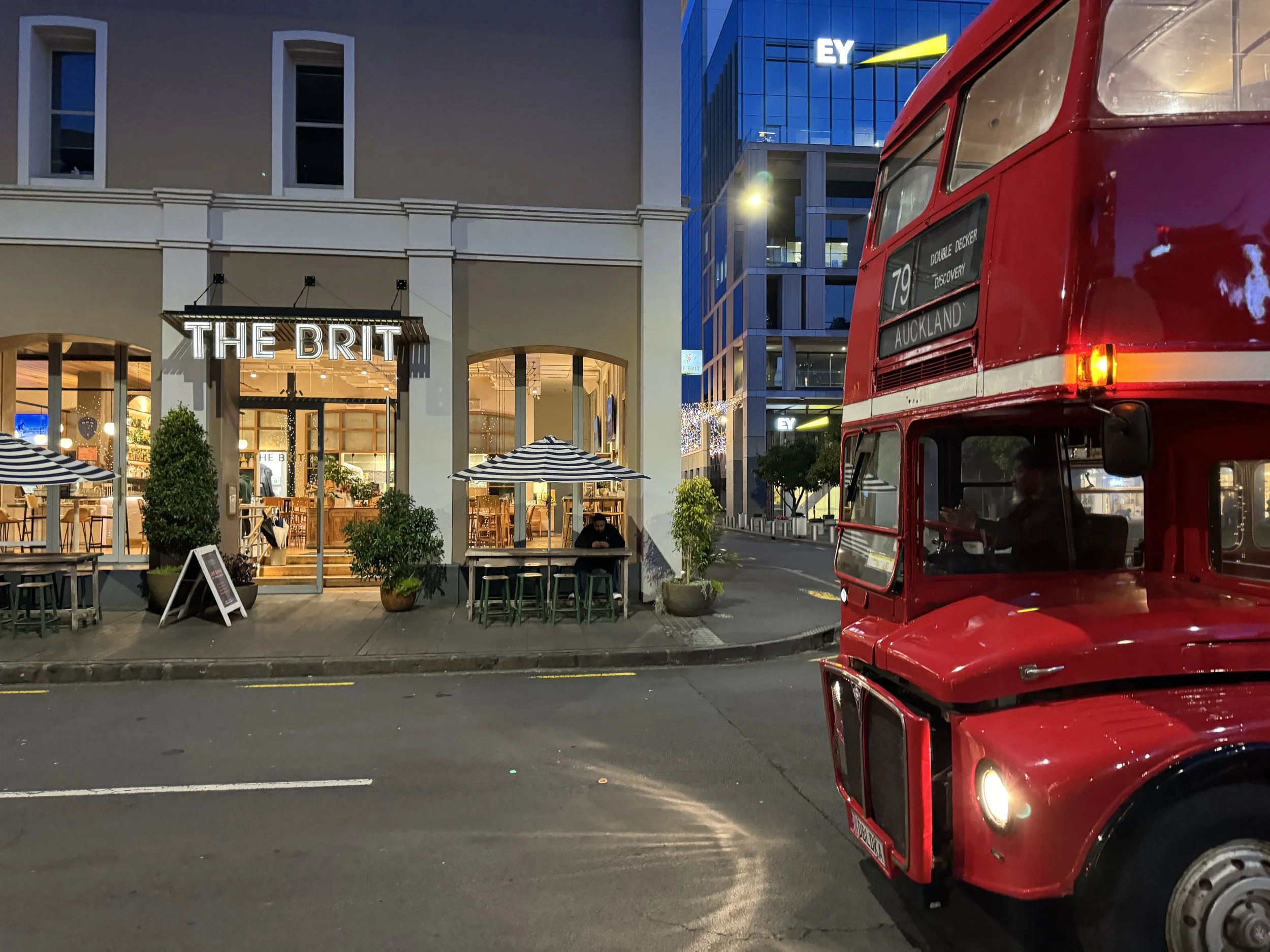 A red double-decker bus parked on the street at night in front of a restaurant named "The Brit" with large windows and outdoor tables, some with striped umbrellas.