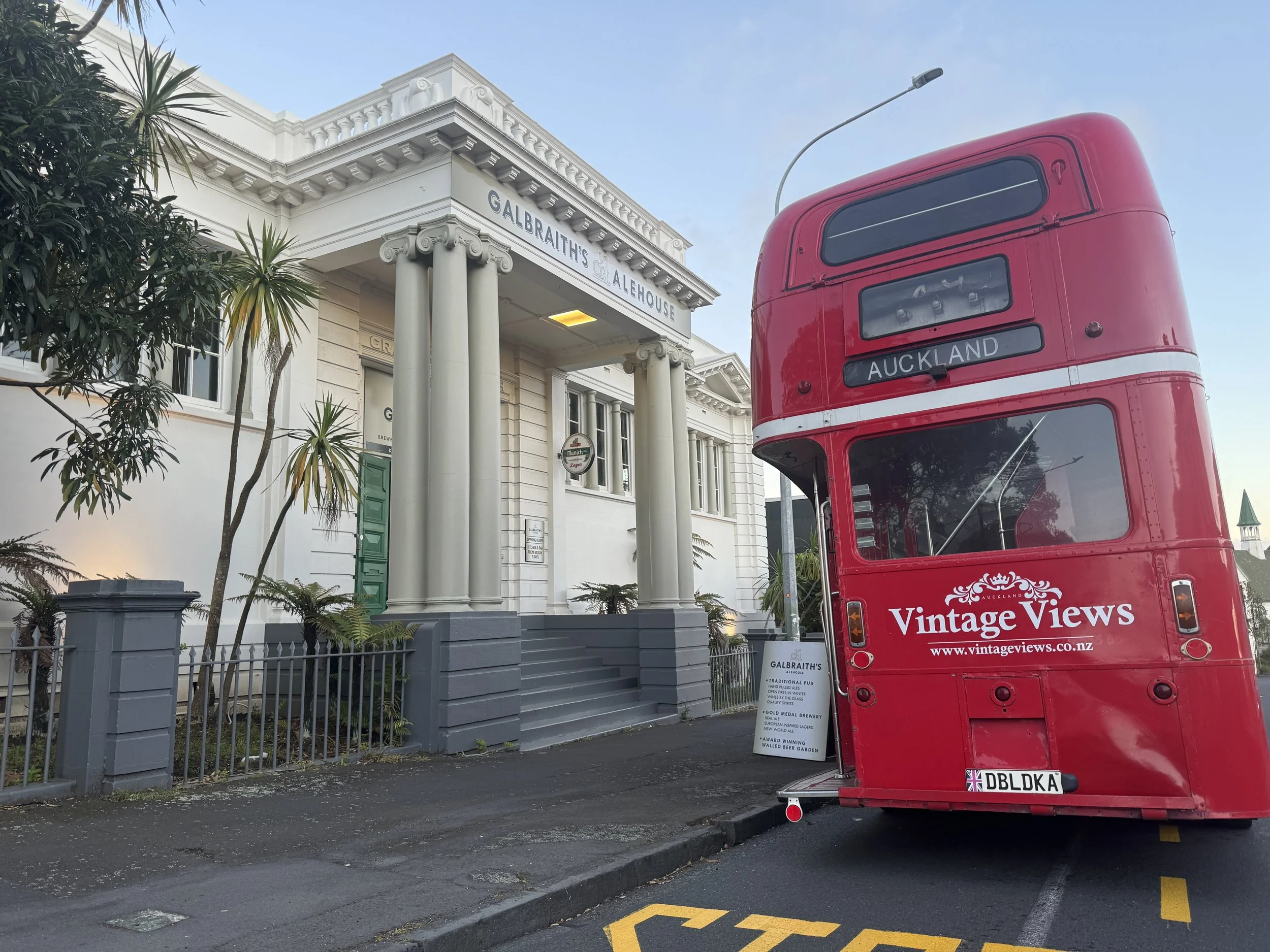A red double-decker bus parked on the street in front of a white building with Greek-style columns and green plants, with a sign indicating a location in Auckland.