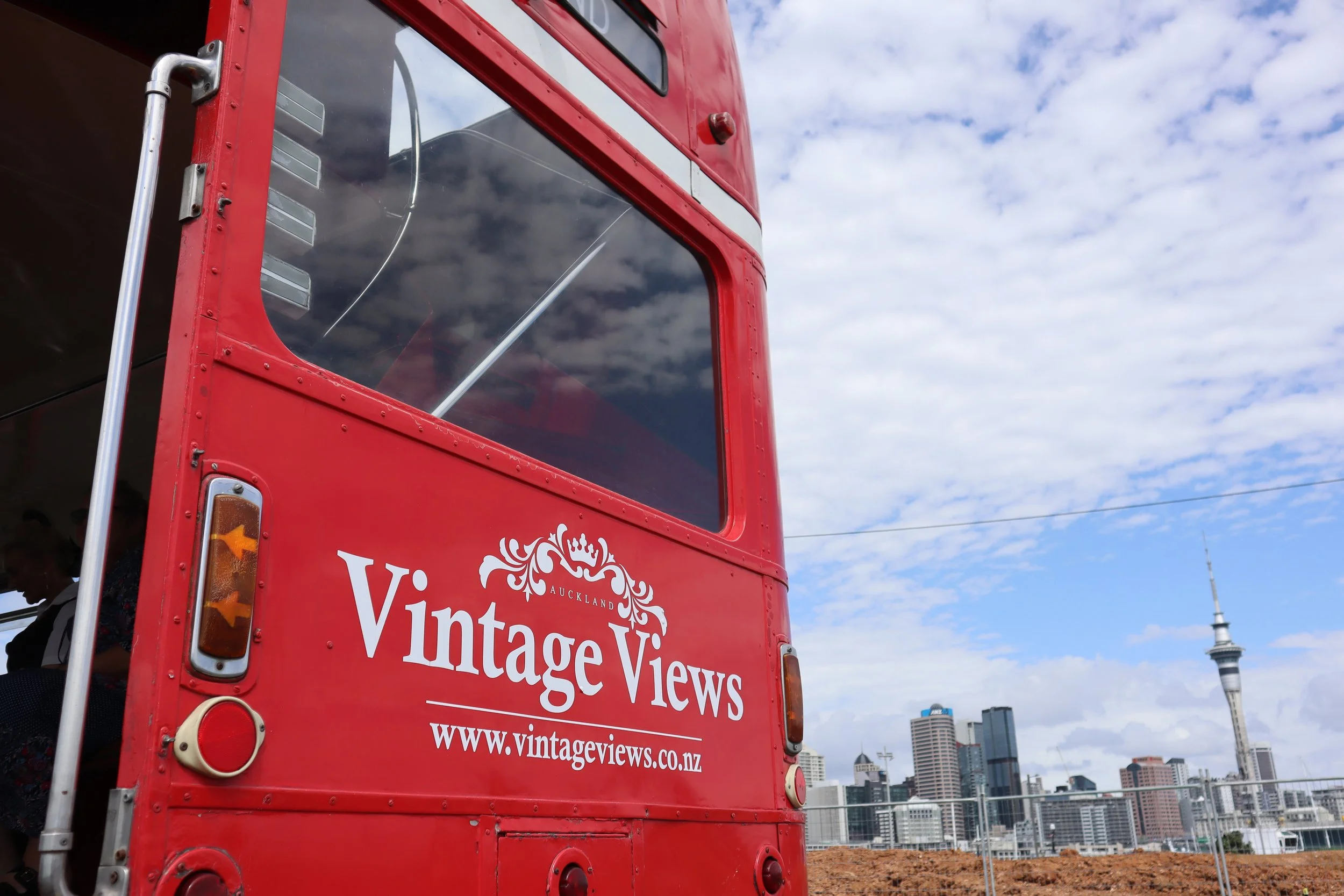 Close-up of the back of a red vintage double-decker bus with Auckland skyline and Sky Tower in the background, showing the sign 'Vintage Views' and website 'www.vintageviews.co.nz'.
