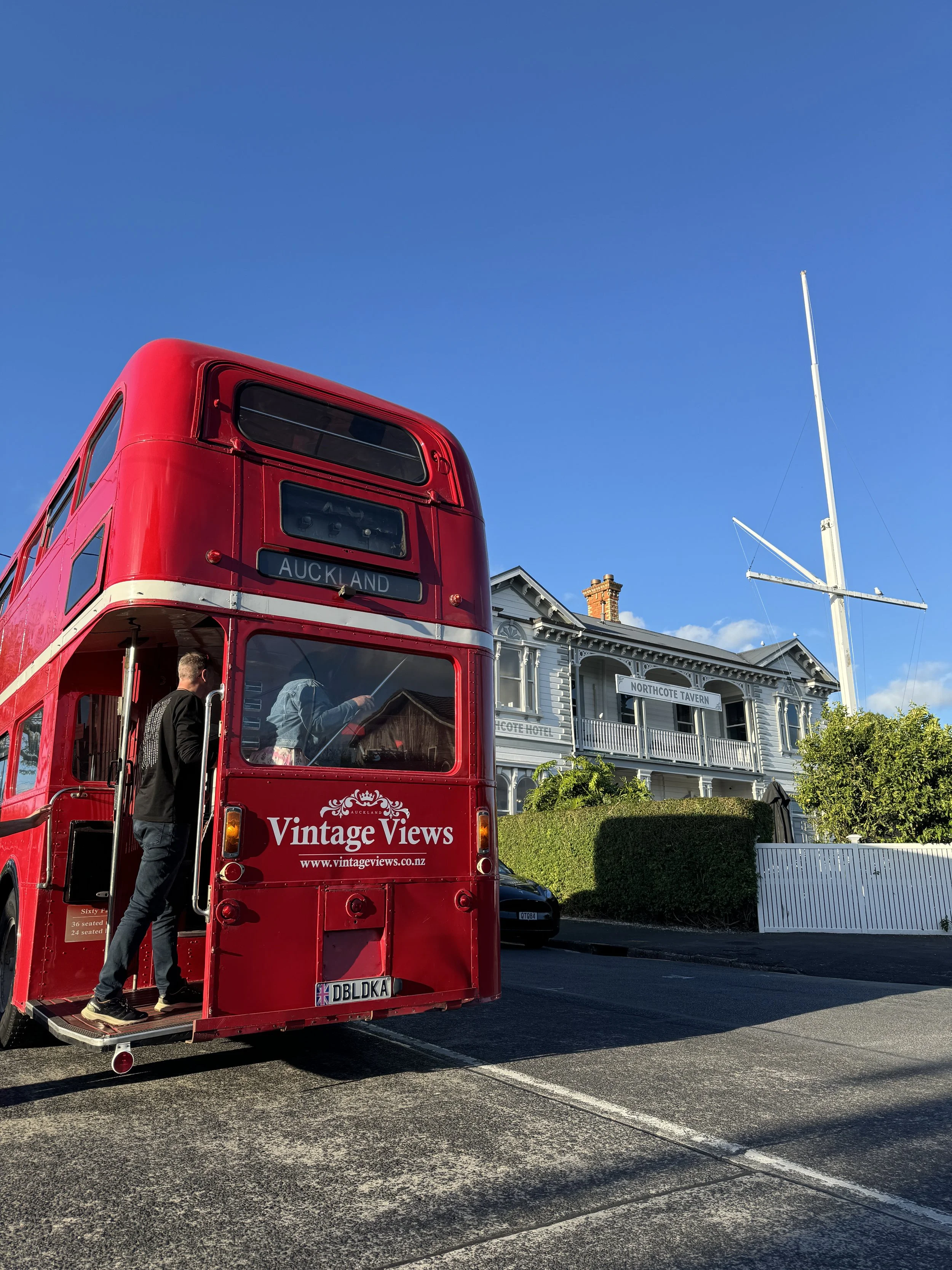 Red vintage double-decker bus with 'Vintage Views' and website on the side, parked on the street near a white wooden building with a sign for Northcote Tavern and Yacht Club.