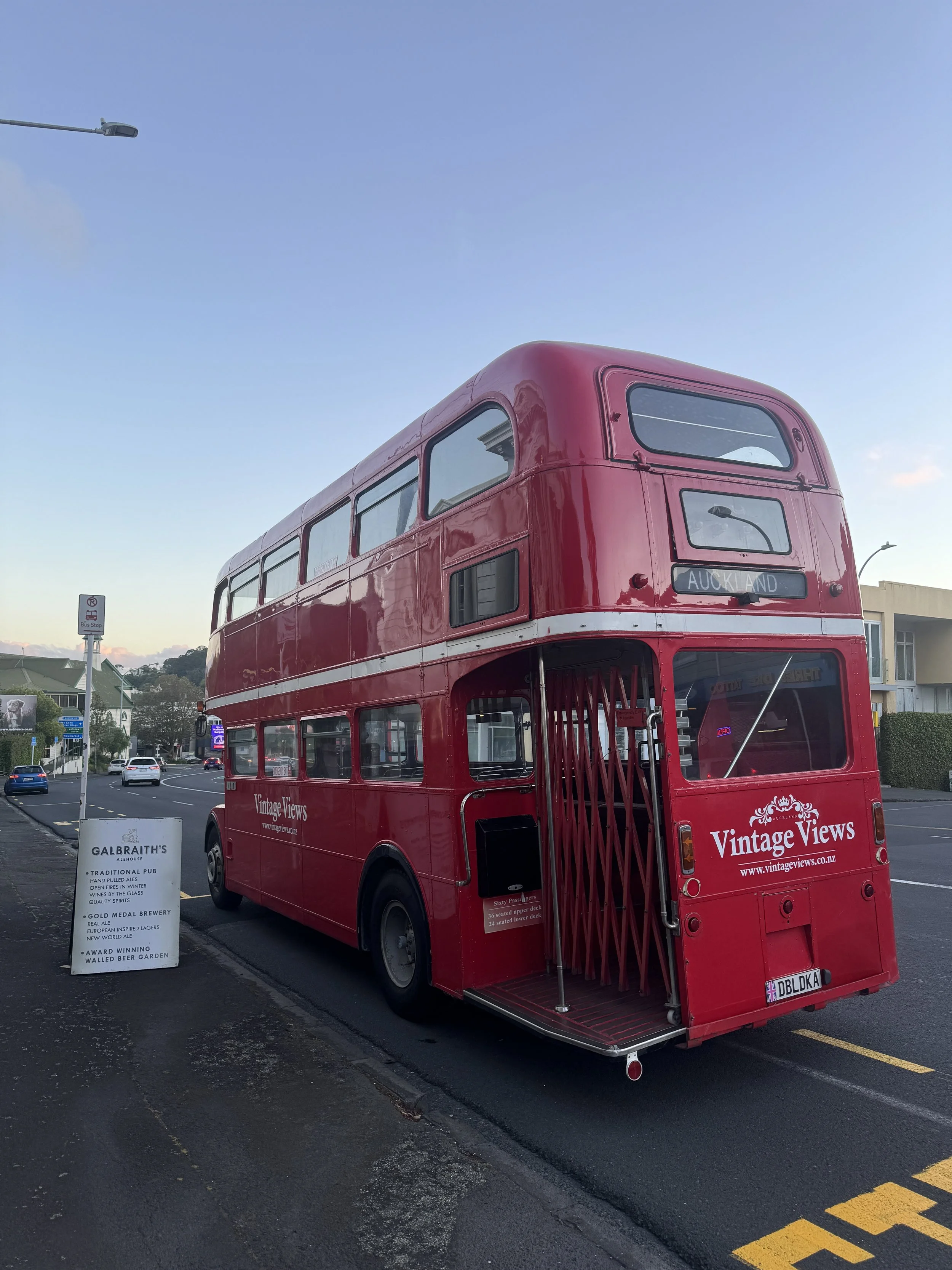 Red vintage double-decker bus with 'Vintage Views' signage parked on street, with a signboard advertising a traditional pub nearby and modern buildings in the background.