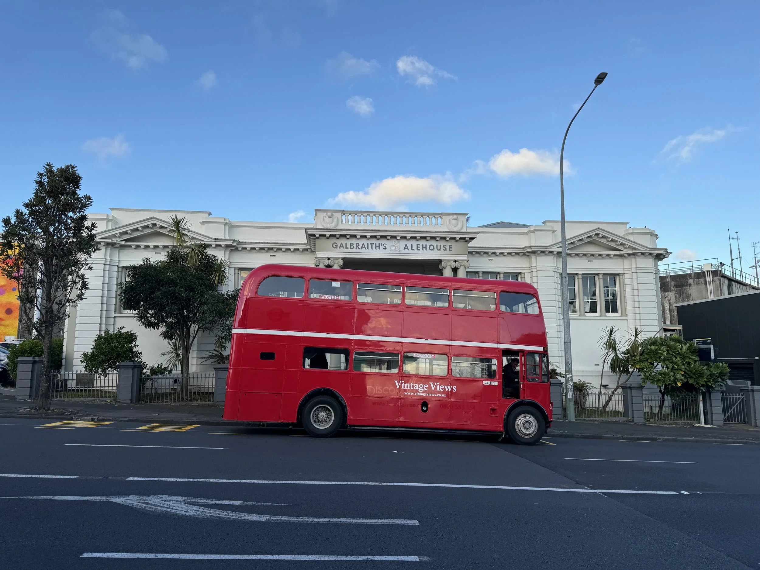 A red double-decker bus parked on the side of a city street in front of a white historic building with a sign reading 'Galbraith's Alehouse.' The sky is partly cloudy with a few trees visible along the sidewalk.