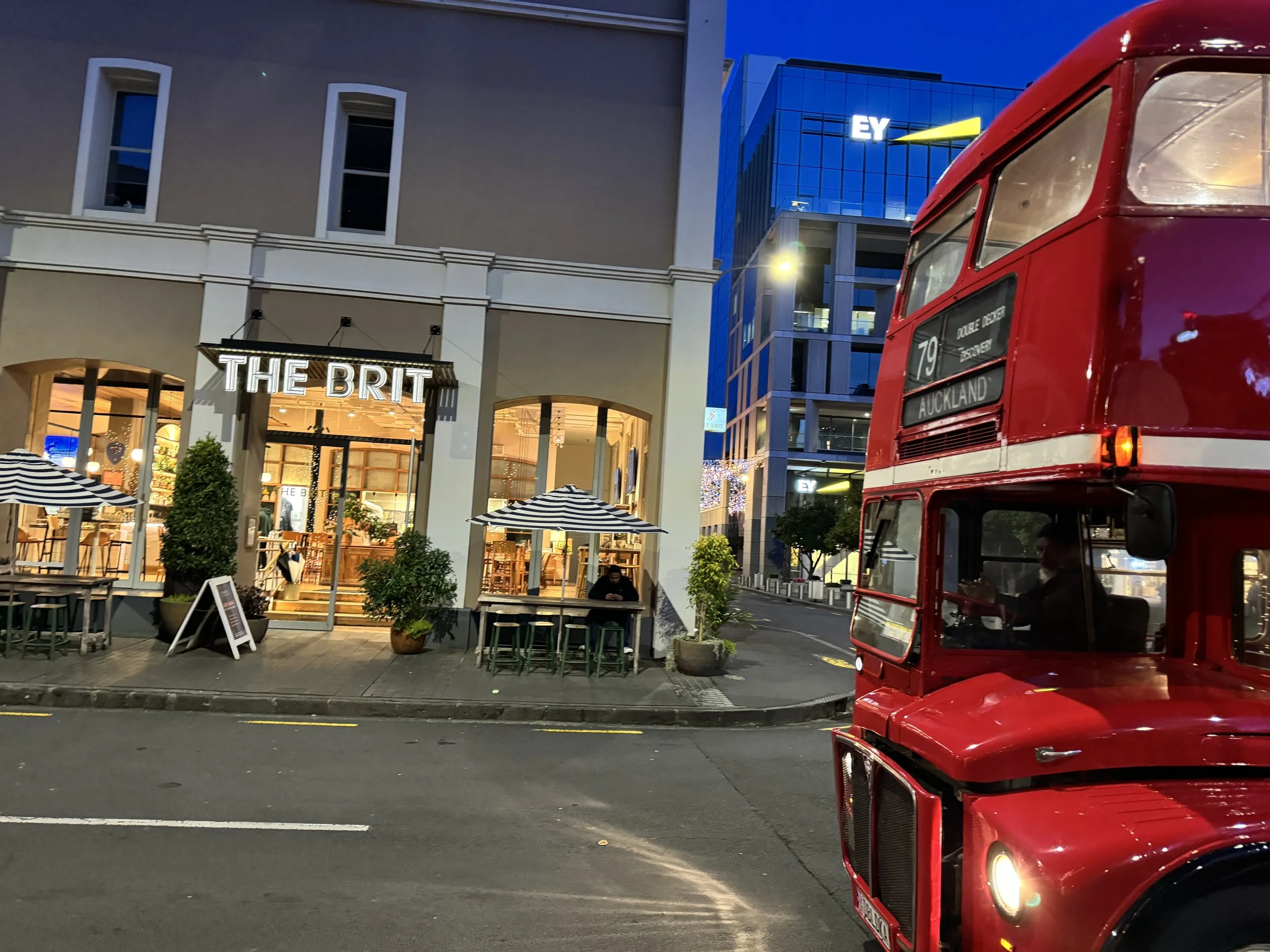 A daytime street scene featuring a modern restaurant called 'The Brit' with outdoor seating and striped umbrellas, and a vintage red double-decker bus with destination signs '79' and 'Auckland' parked on the street.