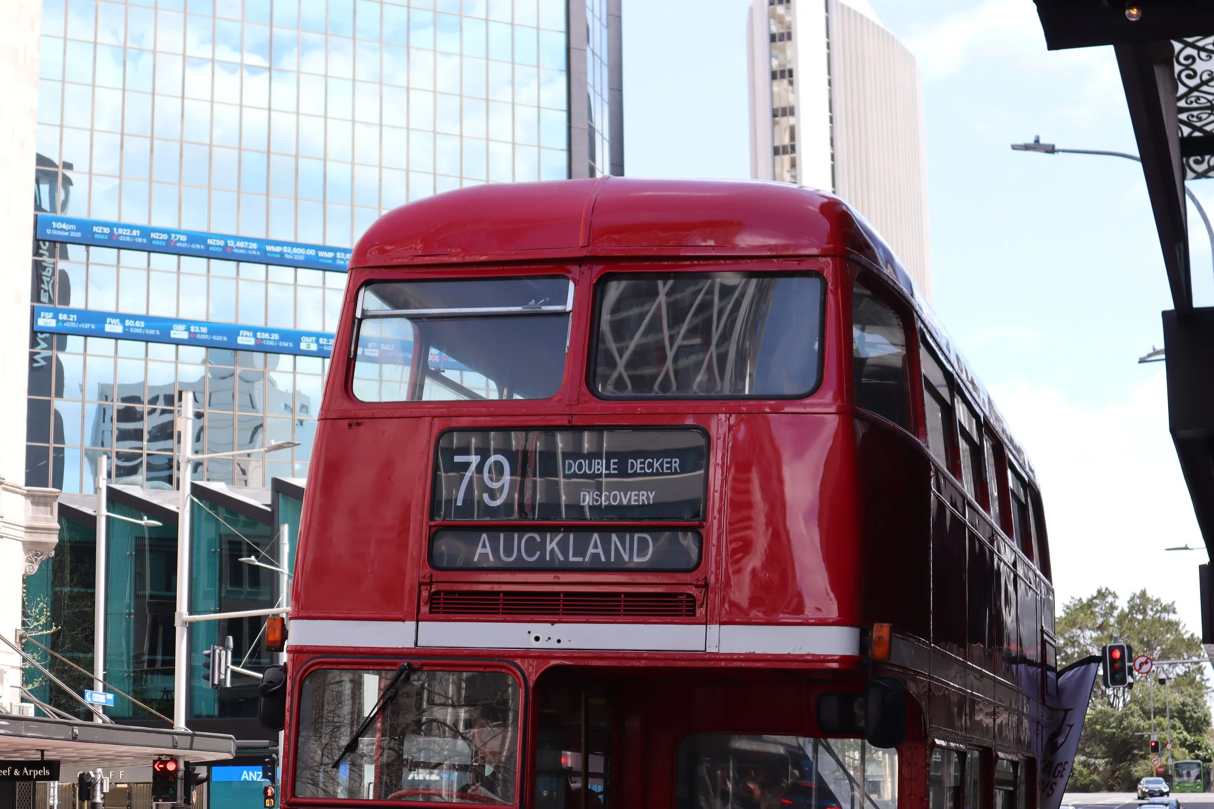Front view of a red double-decker bus showing route number 79 heading to Auckland, with modern glass buildings and a digital stock exchange board in the background.