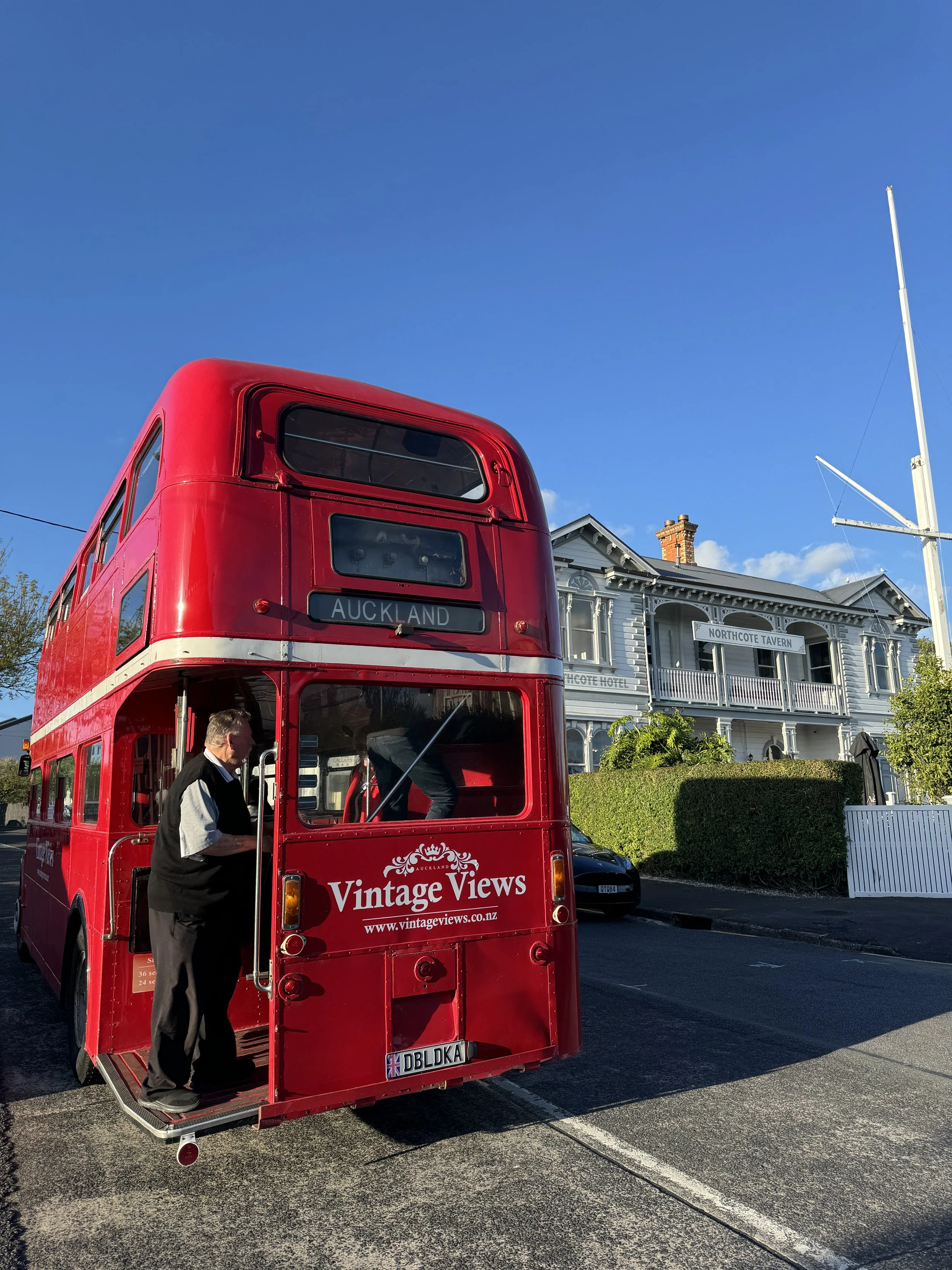 A red double-decker bus with 'Vintage Views' and the website www.vintageviews.co.nz on the back, parked on a street with a man boarding. The bus has signs indicating it’s headed to Auckland. In the background, a building labeled 'Northcote Tavern' an