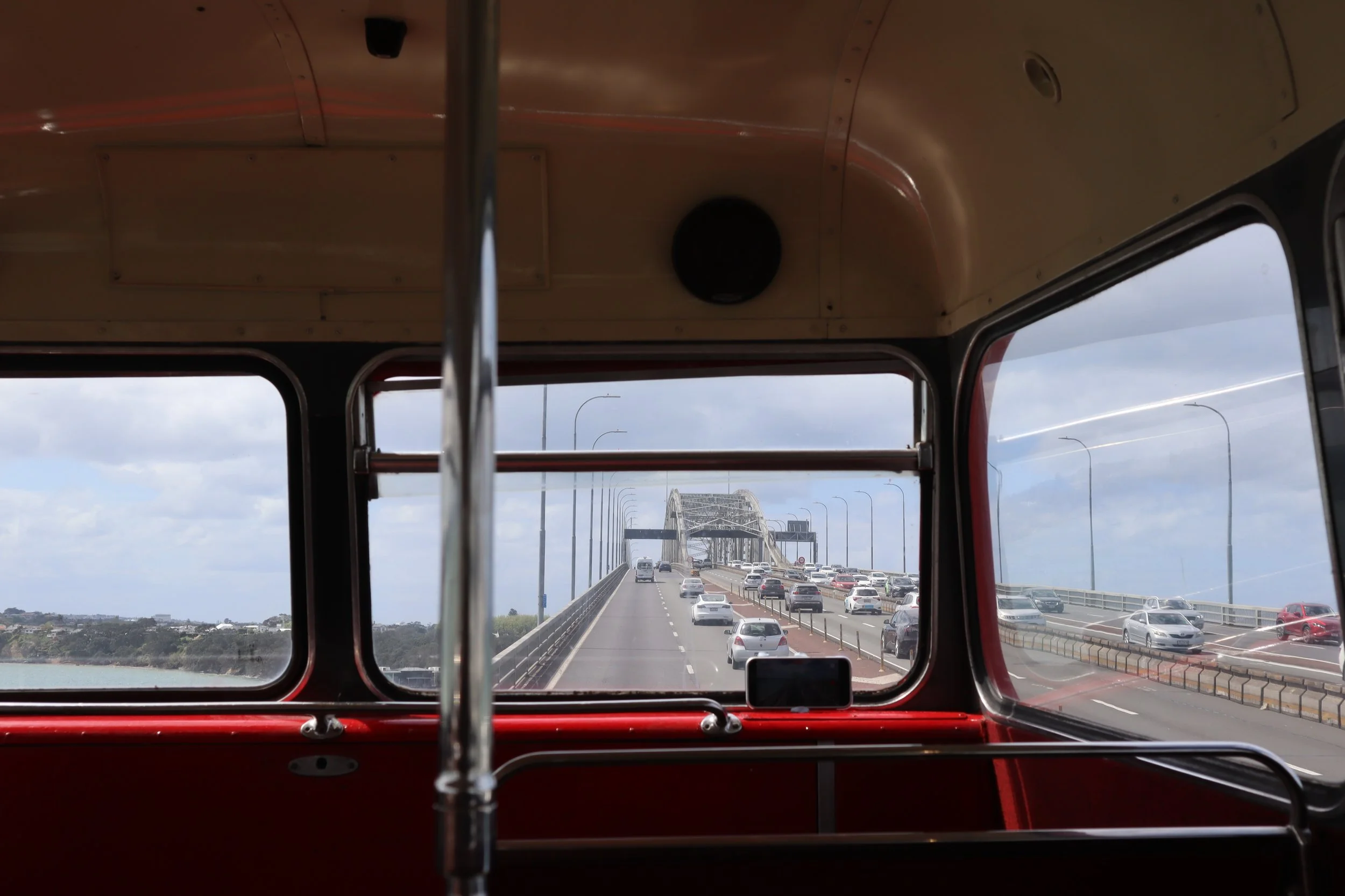 View from inside a bus looking through the front window, showing a bridge with traffic, under a cloudy sky.