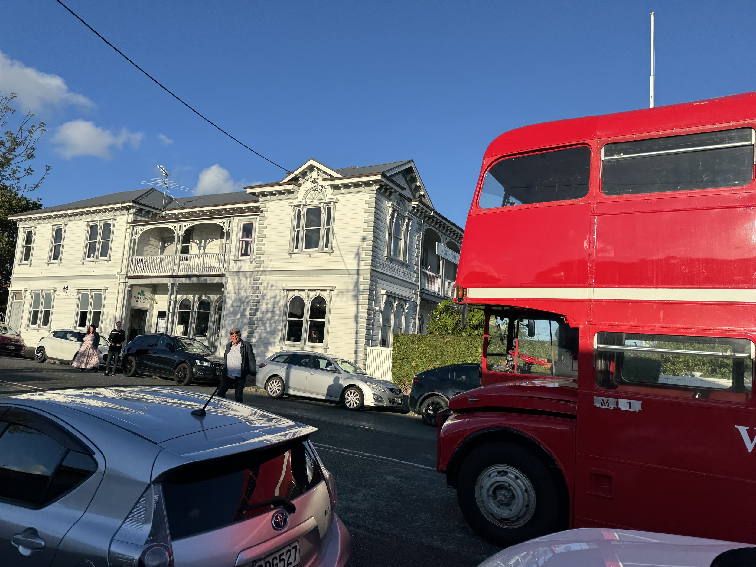 Street scene with several parked cars, a historic white building with bay windows and balconies, and a red double-decker bus on the right side under a blue sky with some clouds.