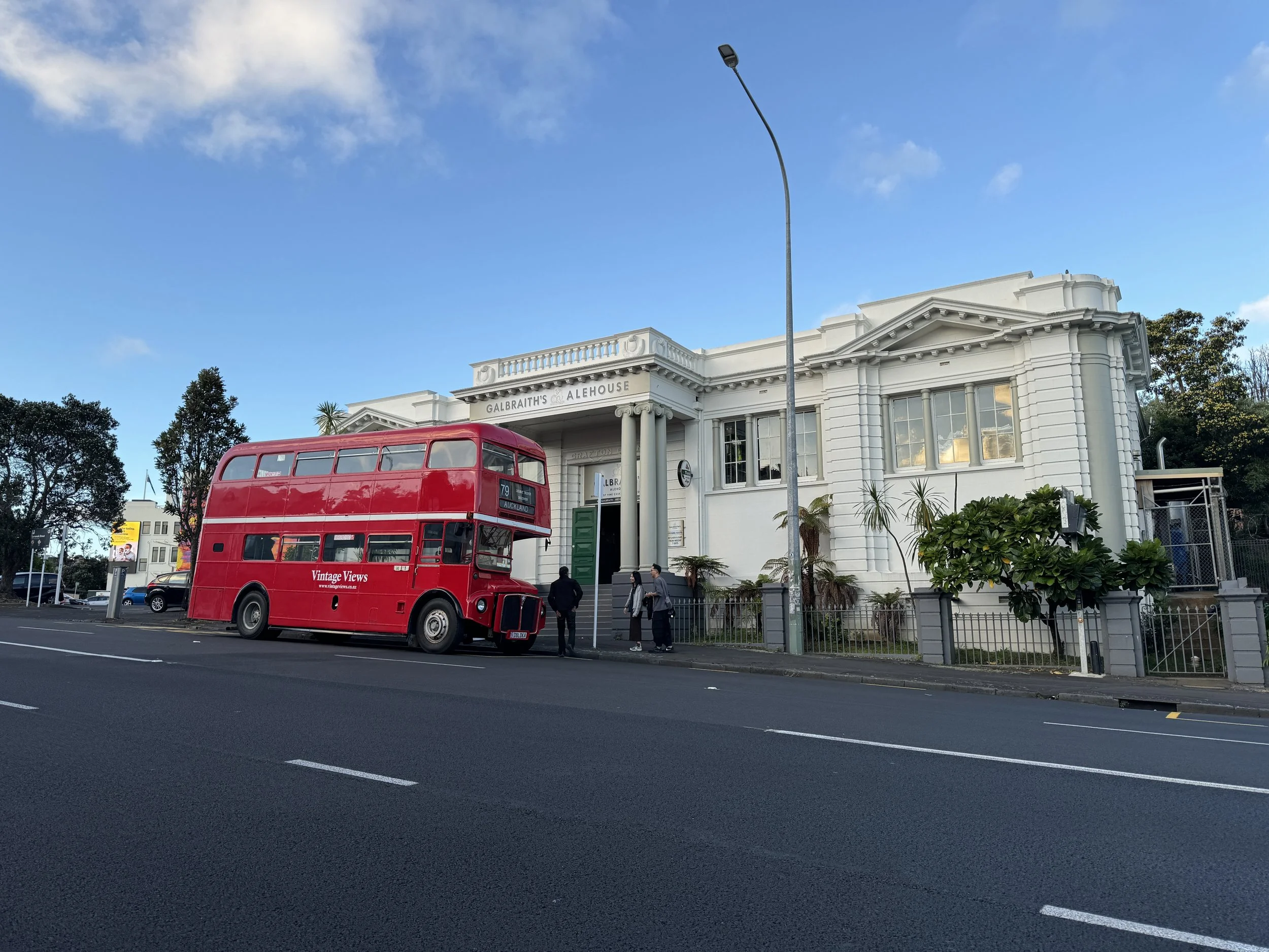 A red double-decker bus parked in front of a white, classical-style building with columns and a sign that reads 'Galbraith's Alehouse'. There are three people standing near the bus, and trees and cars are visible in the background. The sky is mostly 