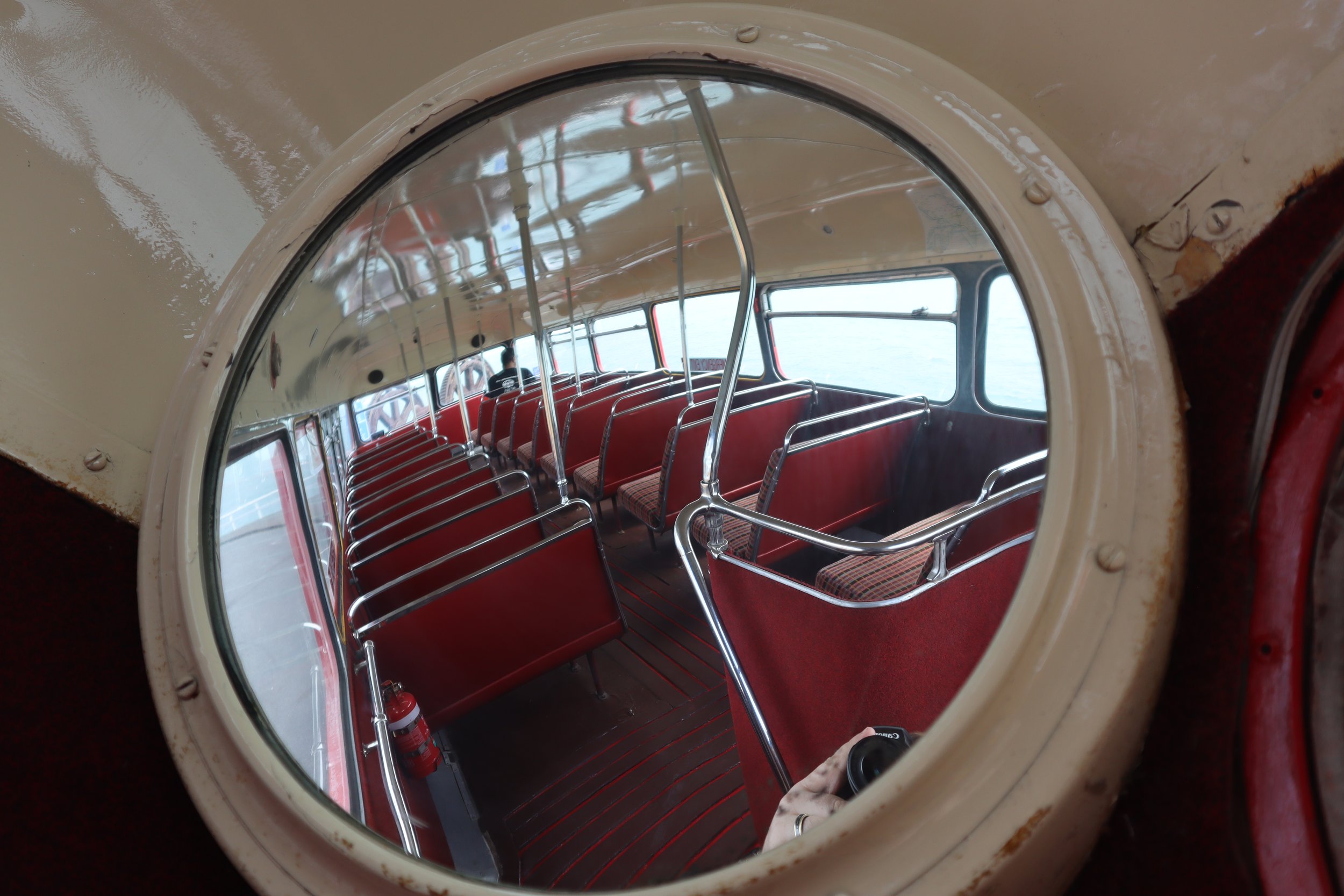 View inside a vintage bus, seen through a porthole window, with rows of red seats and a person in the background.