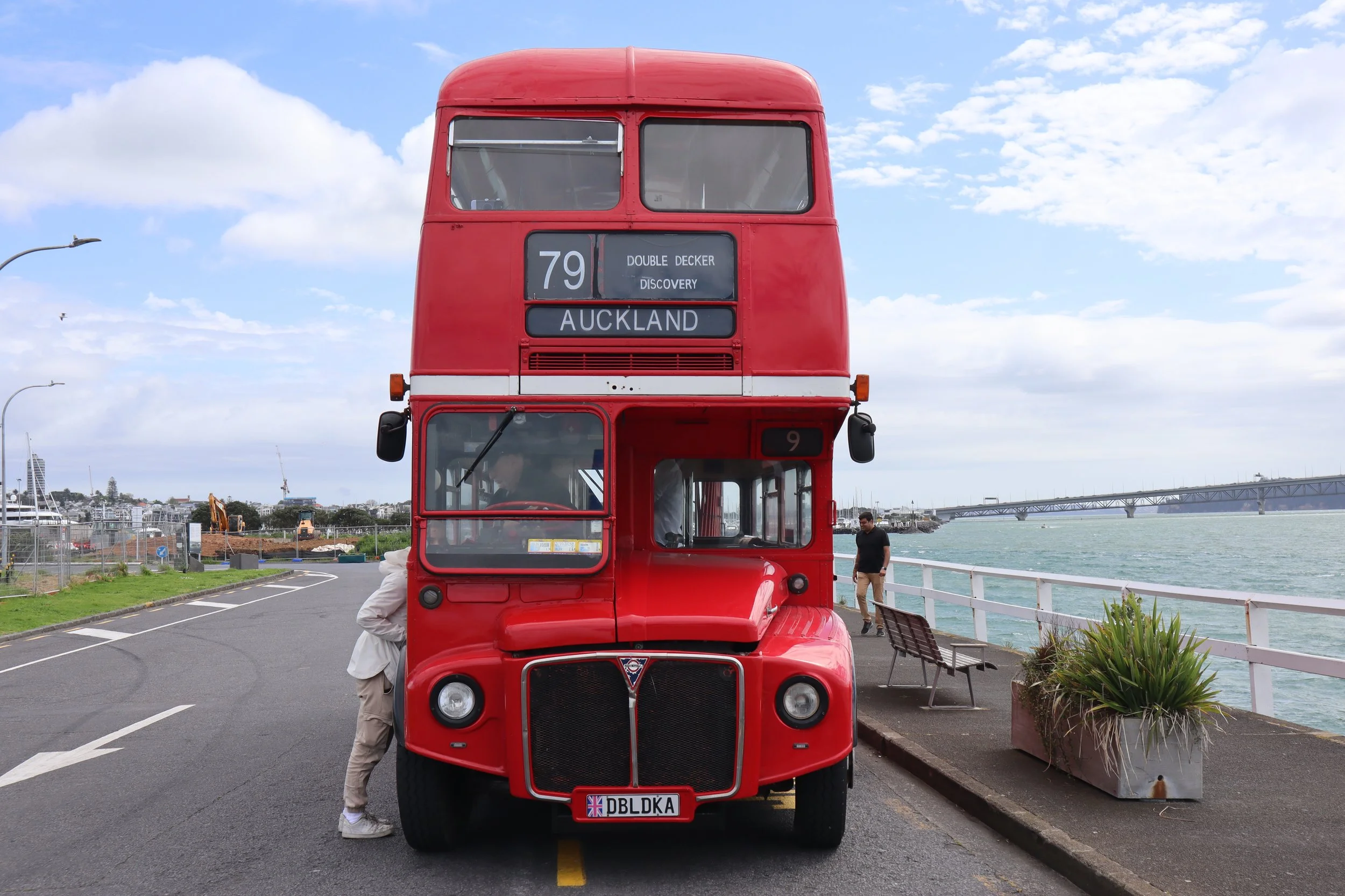 A red double-decker bus parked by a waterfront with a bridge in the background, and people nearby.
