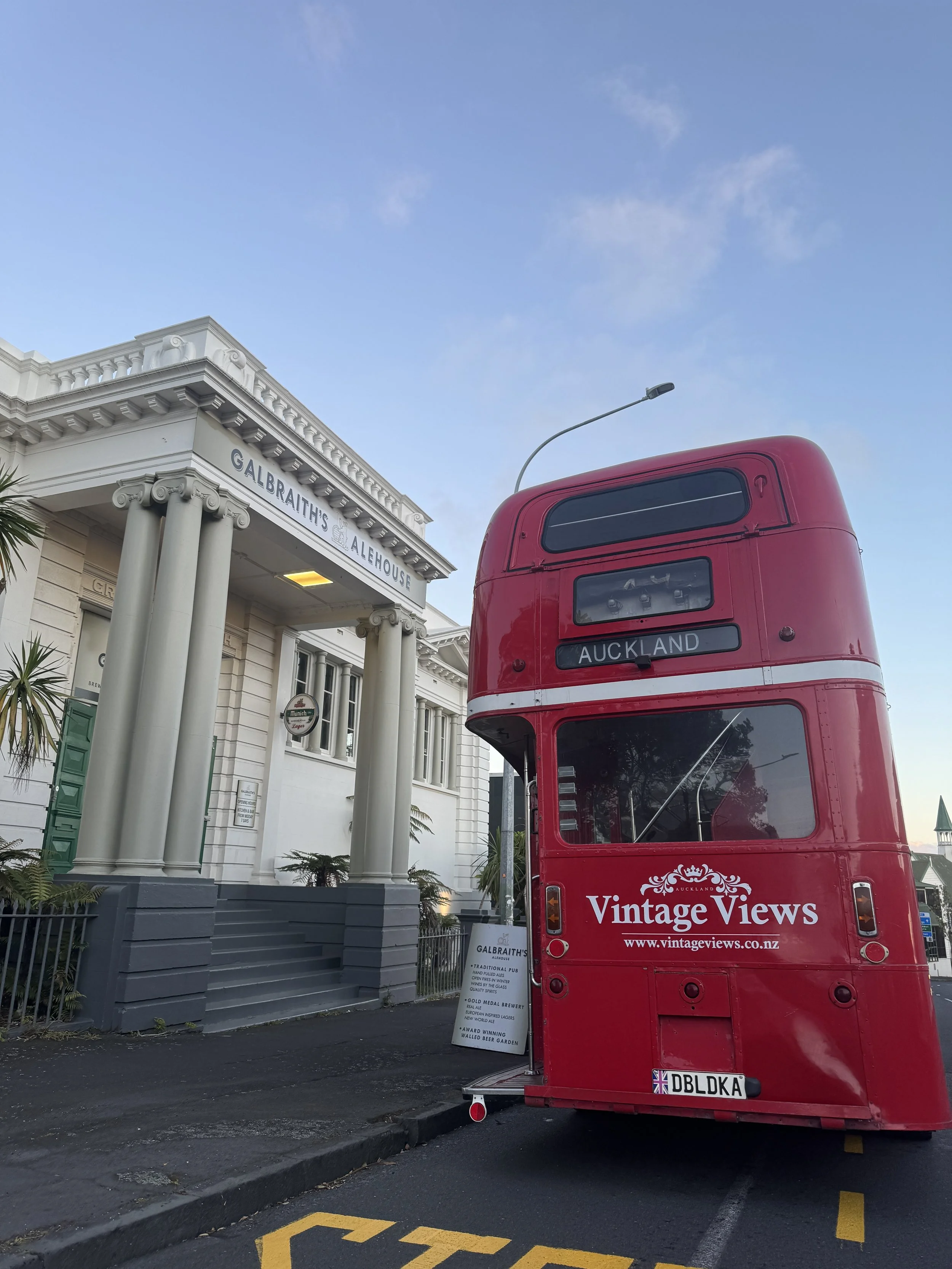 Red double-decker bus with 'Auckland' destination sign and 'Vintage Views' advertisement parked in front of Galbraith's Alehouse, a white classical-style building with columns, on a city street during daytime with a clear sky.
