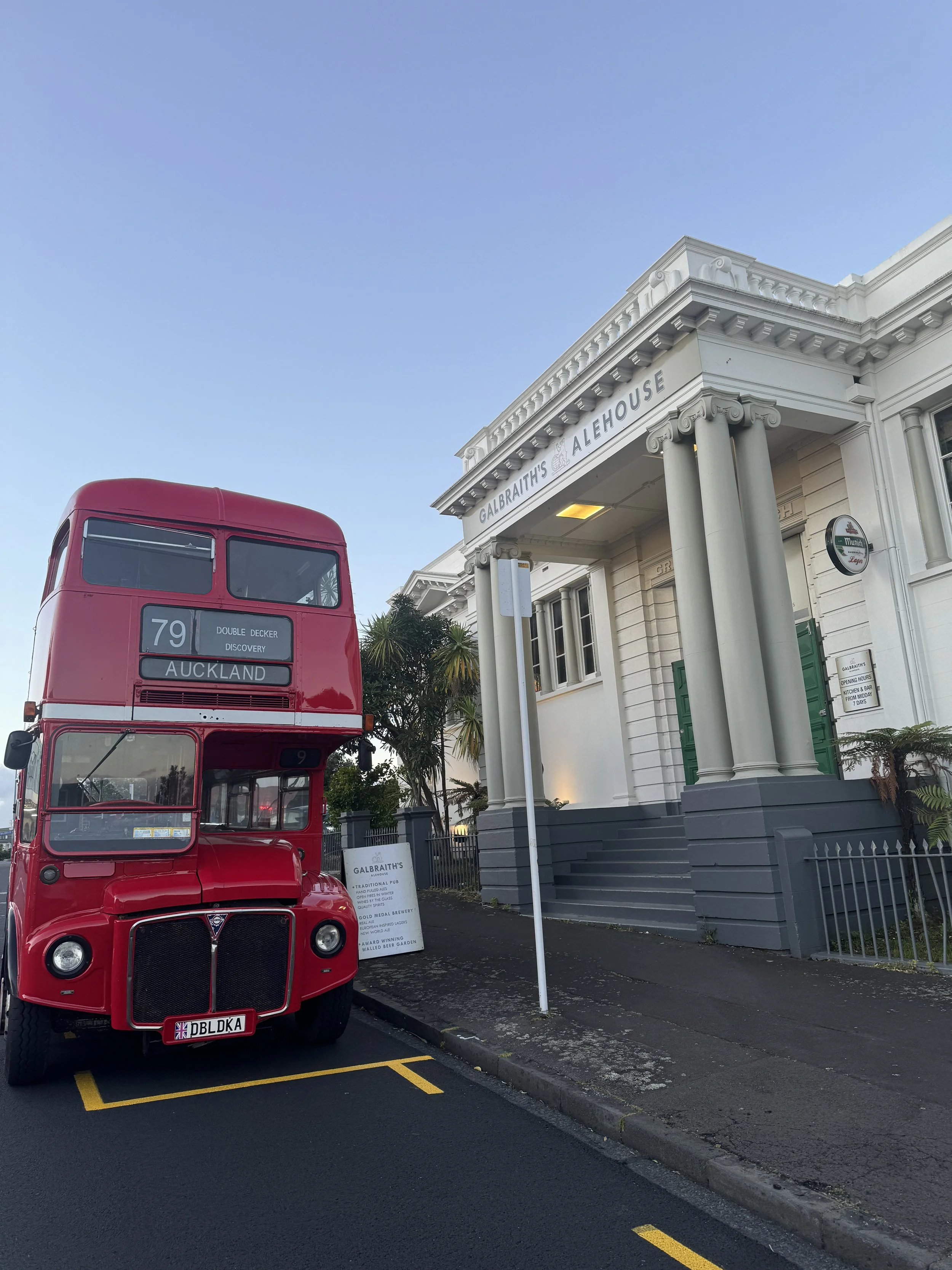 A red double-decker bus parked in front of a white building with columns and a sign that reads 'Galbraith's Alehouse' illuminated at the entrance, with a clear blue sky overhead.