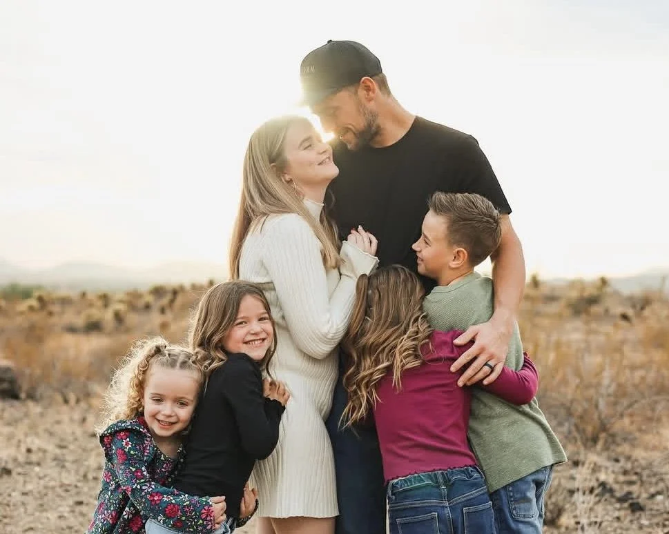 A happy group of six people, including two adults and four children, embracing outdoors with a sunset in the background, smiling and enjoying each other's company.