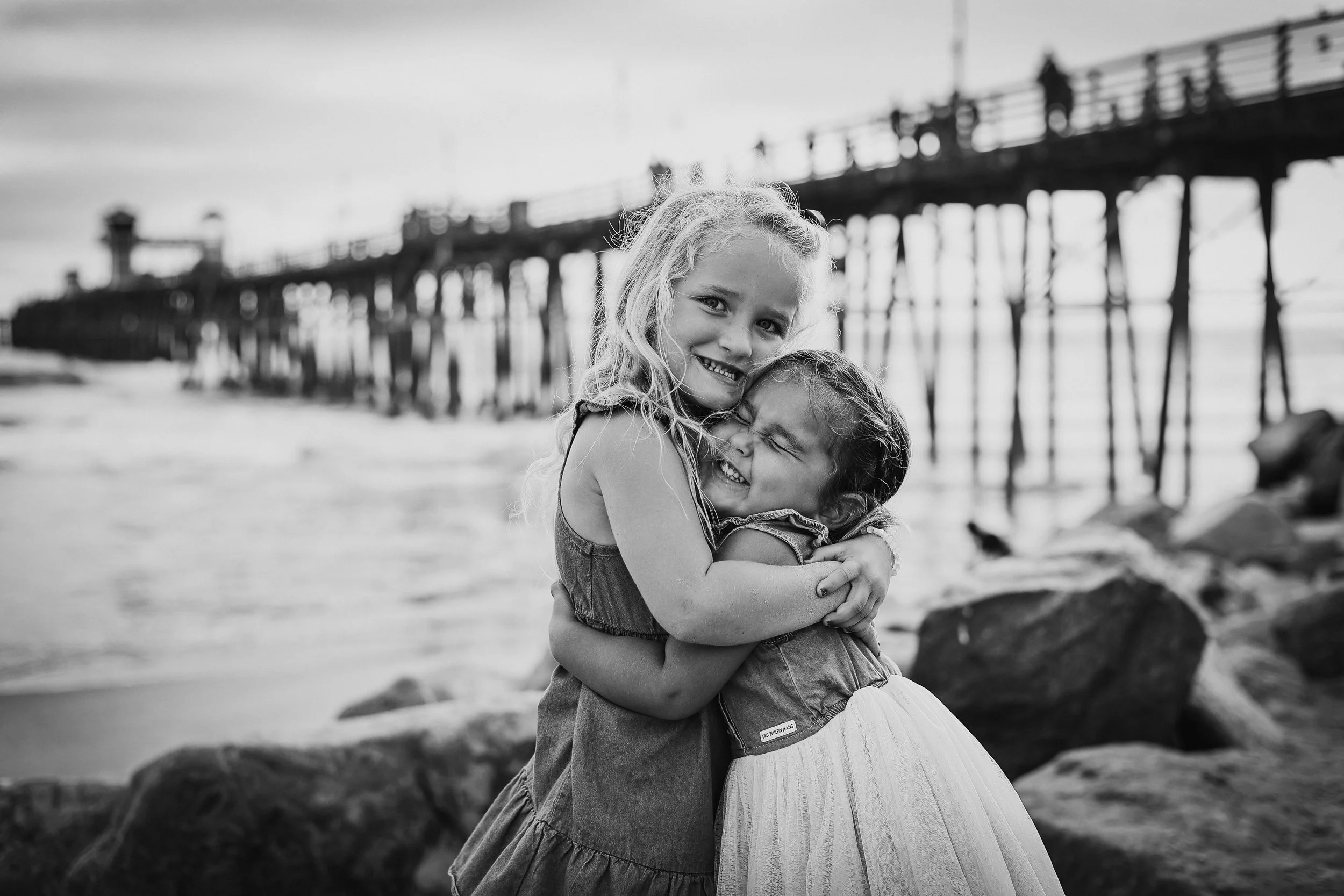 Two young girls hugging on a beach with a pier in the background, smiling and enjoying each other's company.