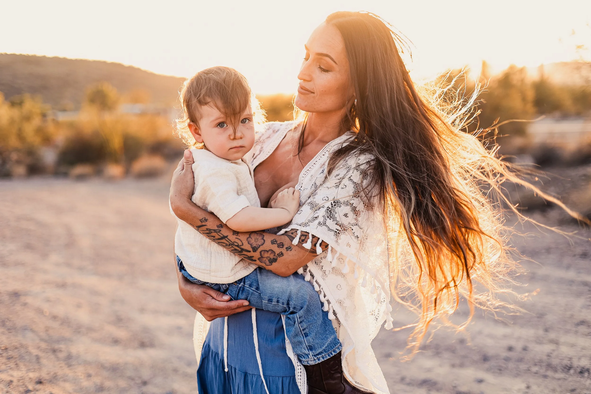 A woman with long, wavy hair holding a young child with short hair outdoors at sunset.