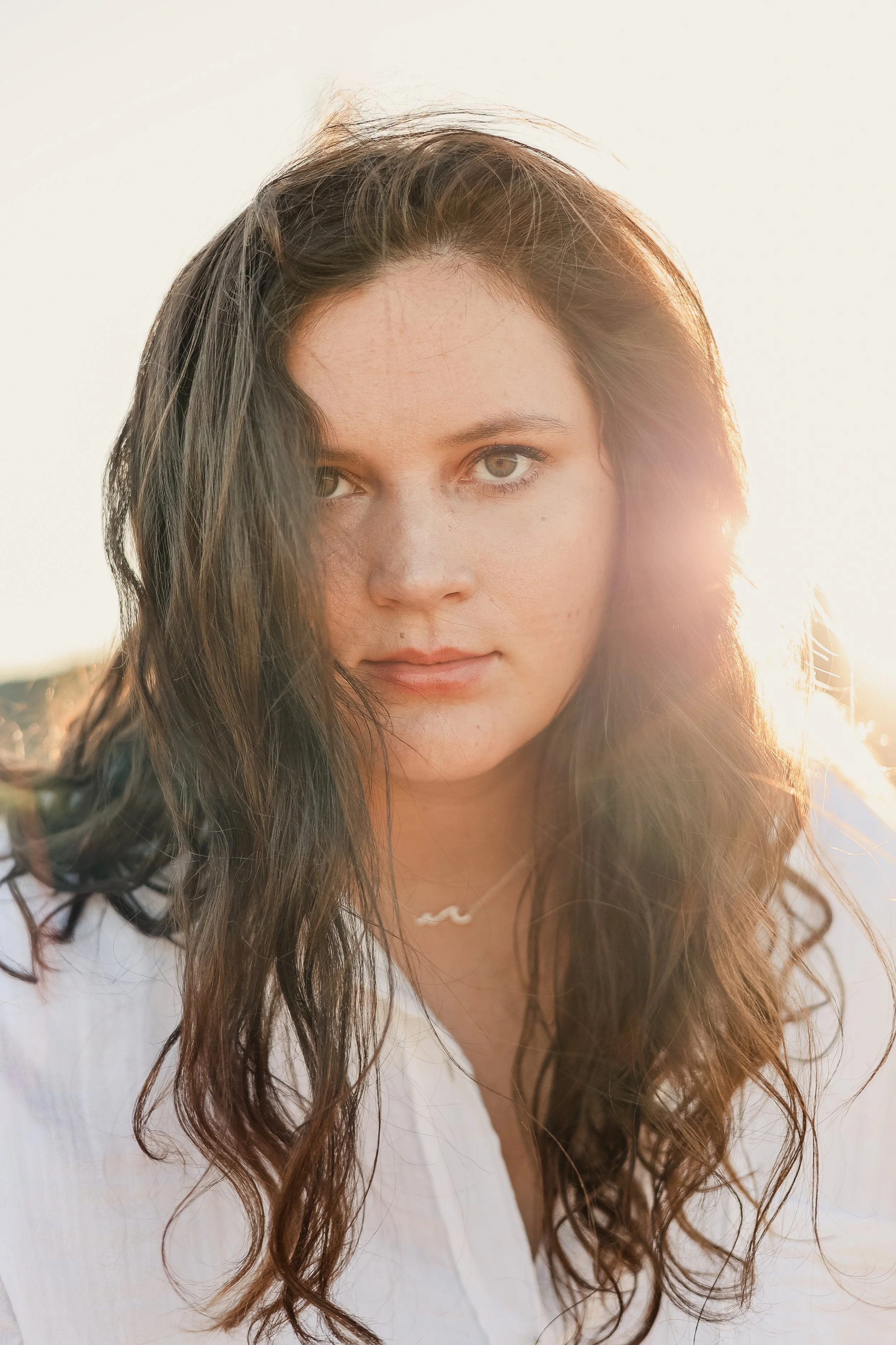 A young woman with long, dark, wavy hair and light skin, wearing a white top, with sunlight behind her, looking into the camera.