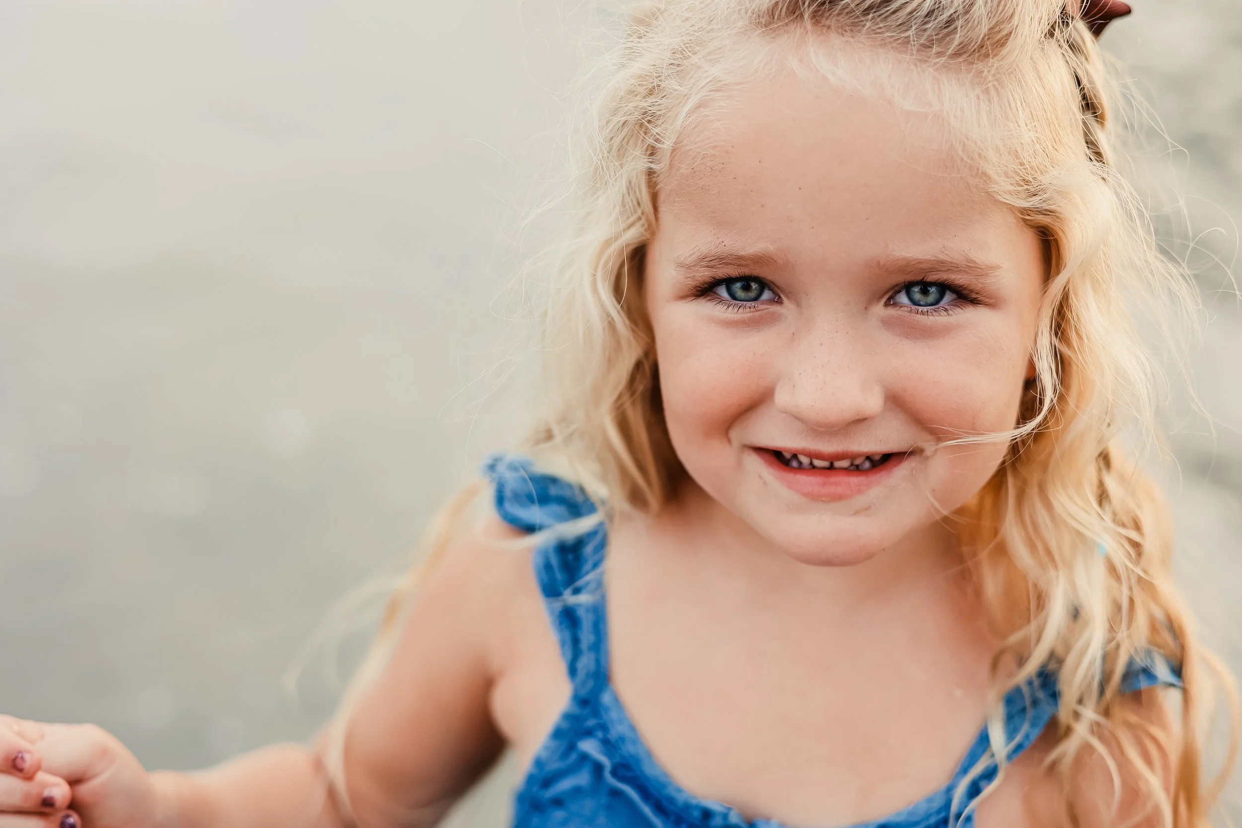 Close-up of a young girl with long blonde hair, blue eyes, and freckles, smiling while at the beach during daytime.