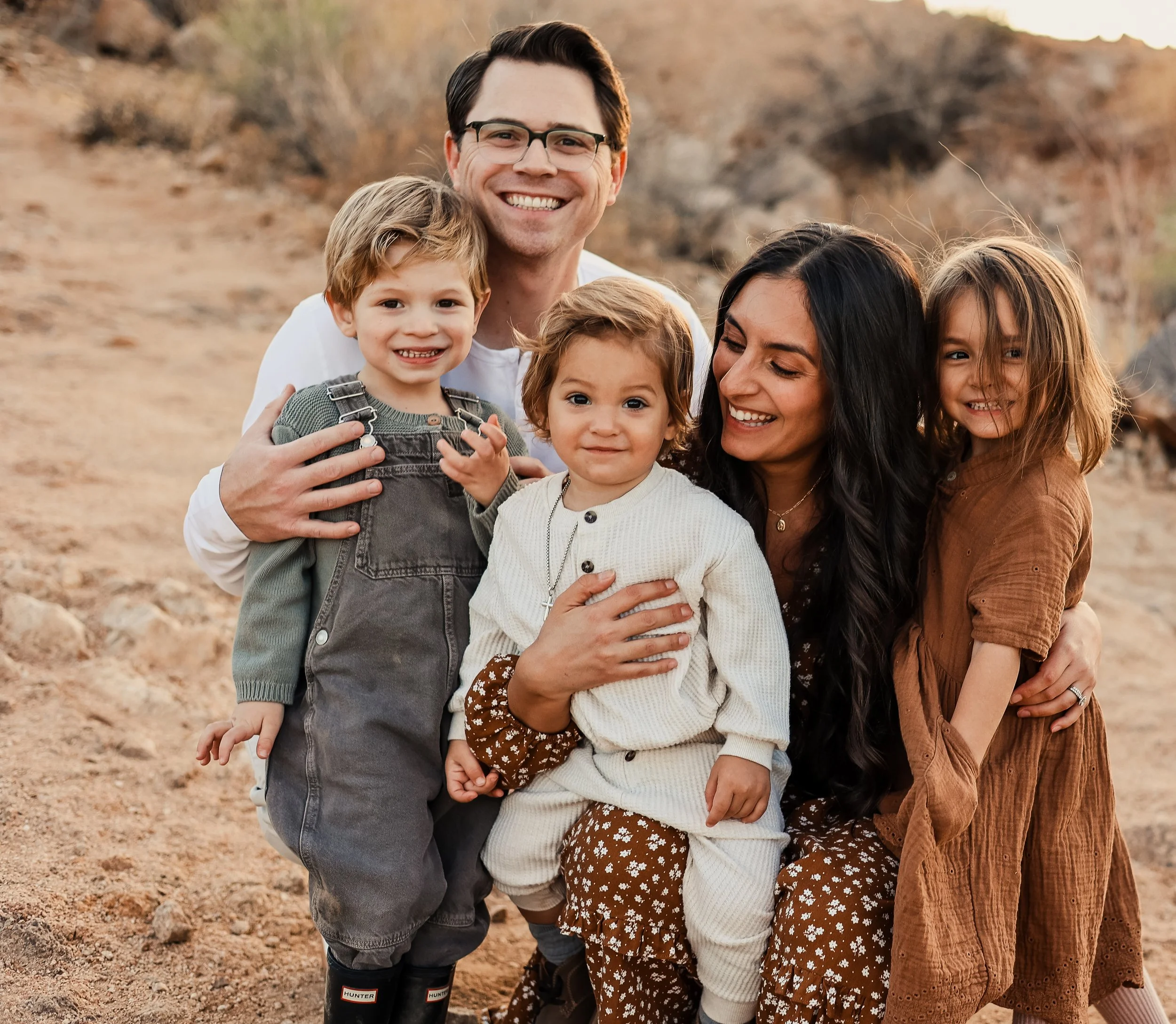 A happy family of six members, including two adults and four children, outdoors in a desert-like environment during sunset, all smiling and embracing each other.