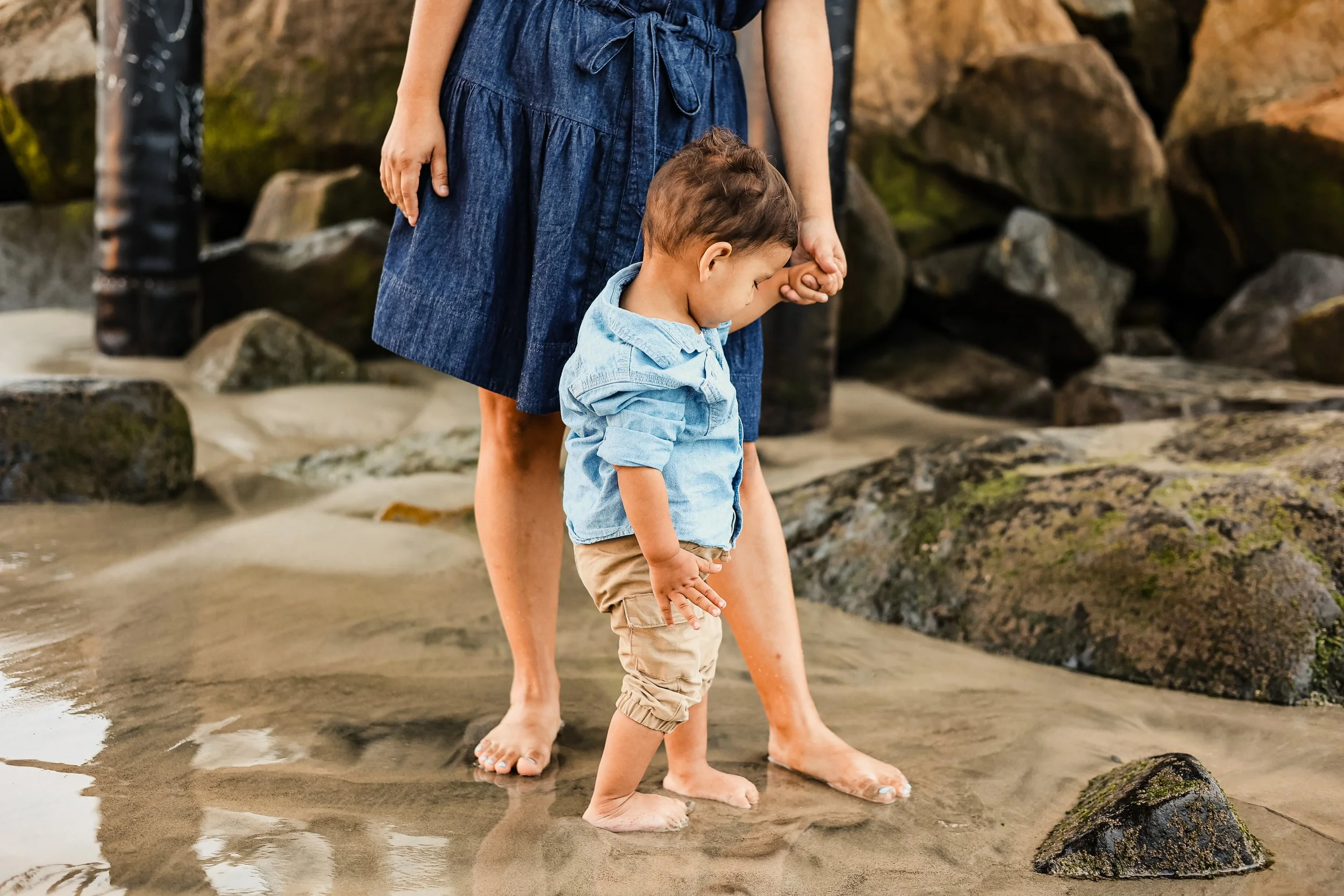 A woman and a young boy standing barefoot in shallow water on a sandy beach with rocks in the background, holding hands.