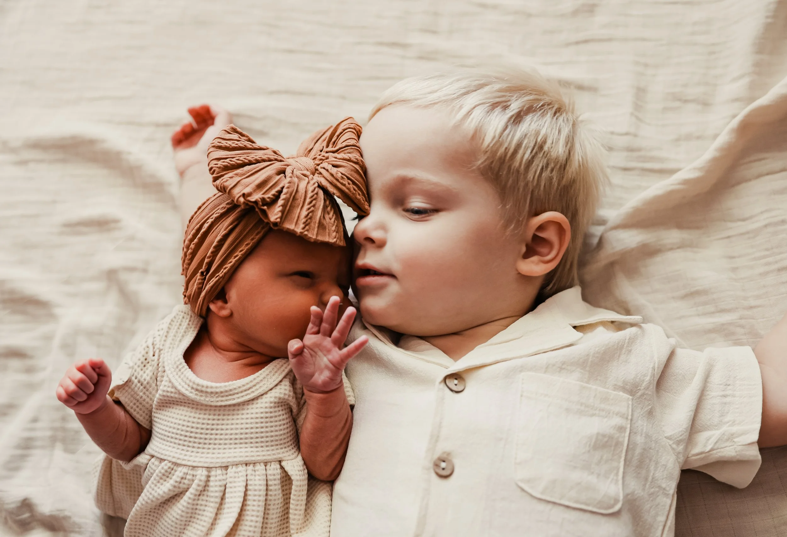 An adorable young blonde boy lying on a bed, gently touching a tiny newborn baby girl with a headscarf, both resting on a soft, white sheet.