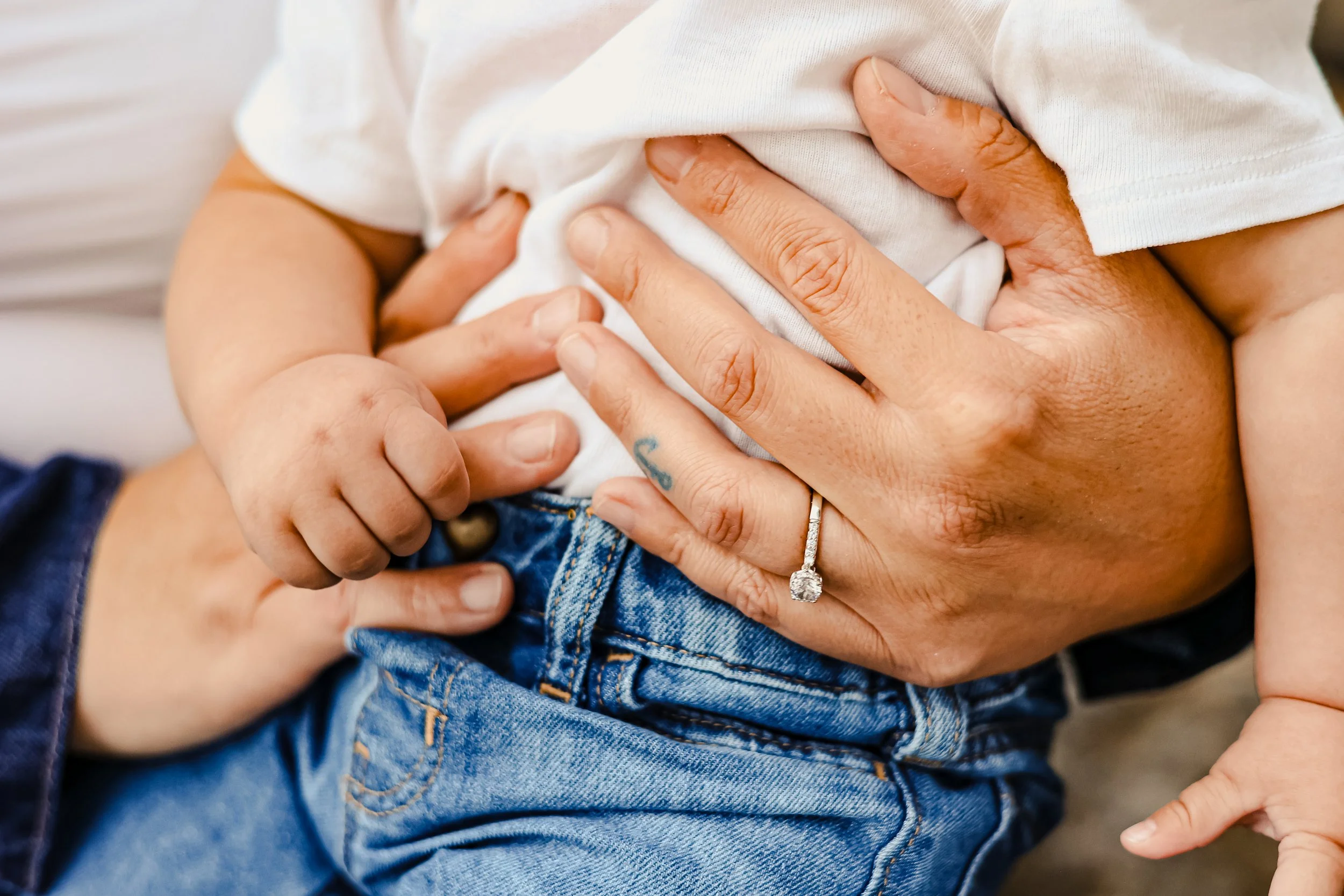 Close-up of a hand with an engagement ring holding a child's hand onto a person's abdomen, both wearing white and blue clothing.