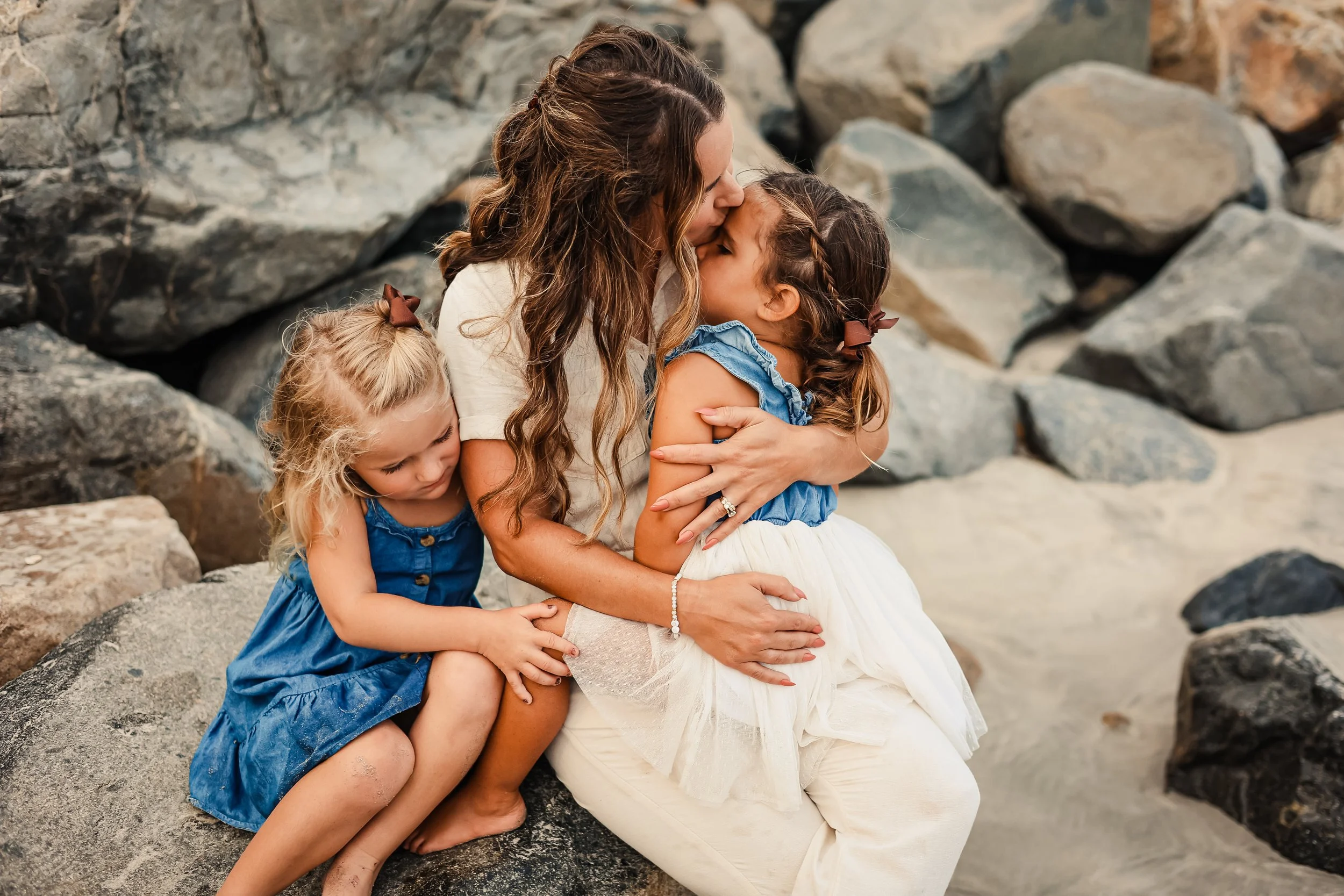 A woman sitting on rocks at the beach hugging and kissing two young girls wearing blue dresses.