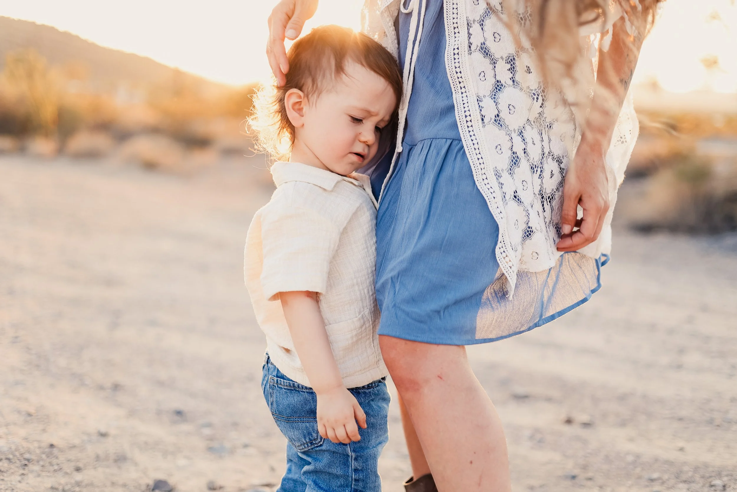A young boy is holding his stomach, looking distressed, while standing next to a woman on a sandy terrain during sunset. The woman is gently touching the boy's head.