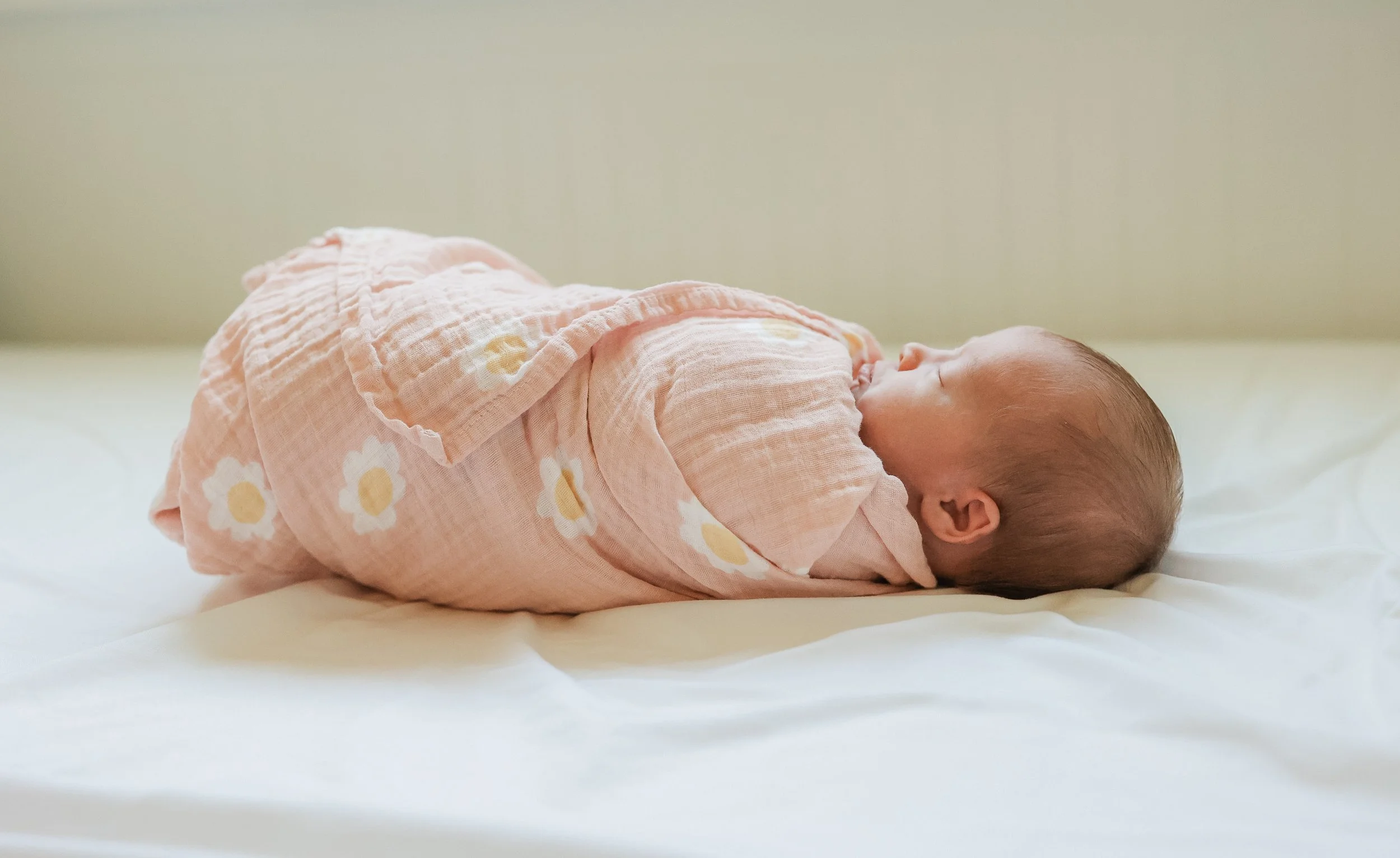 A newborn baby wrapped in a pink blanket with white and yellow daisy patterns, lying on a white surface.