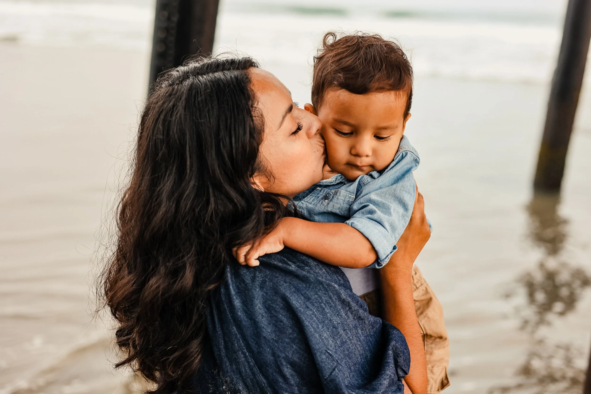 A woman is holding and kissing a young boy on a pier by the water.