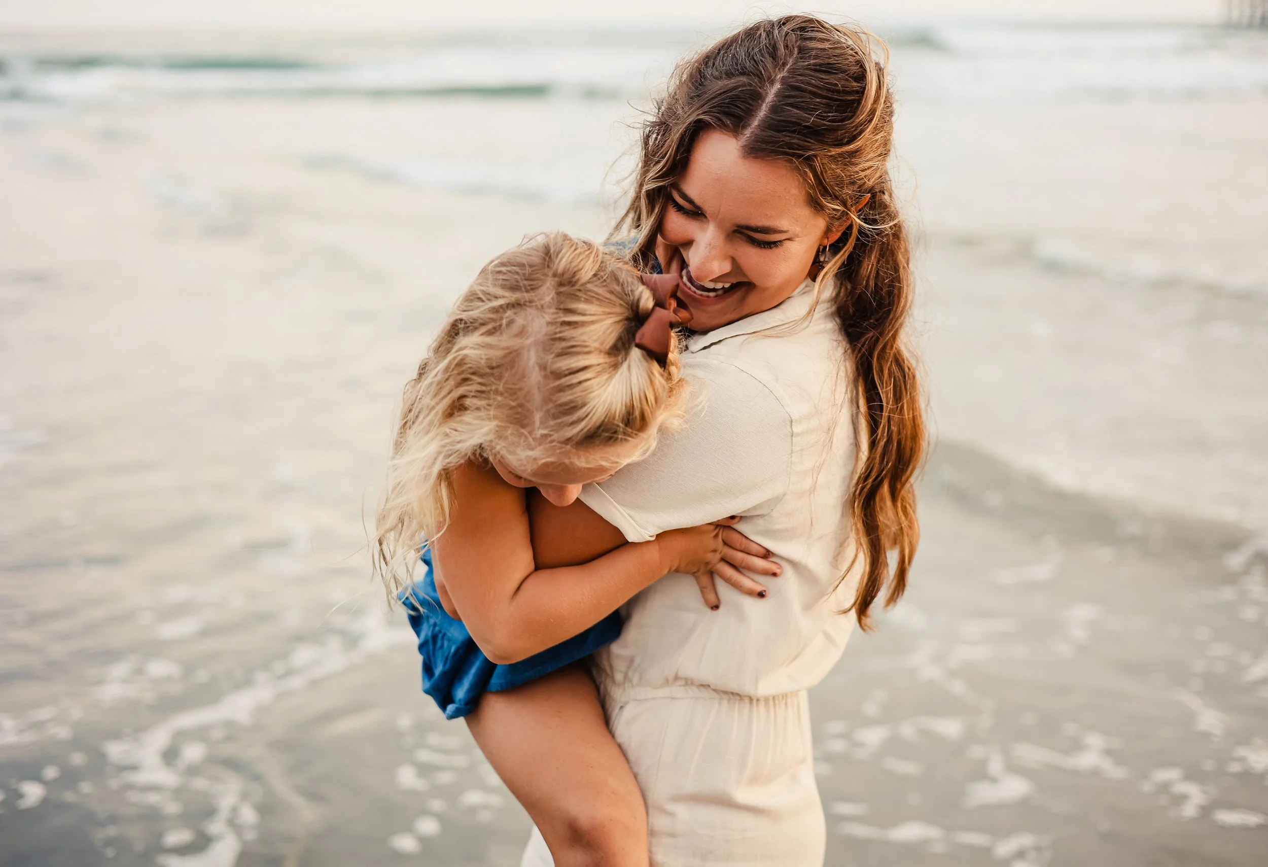 A woman with long brown hair holds a young girl with blonde hair and a brown hair bow, both smiling and hugging at the beach with ocean waves in the background.