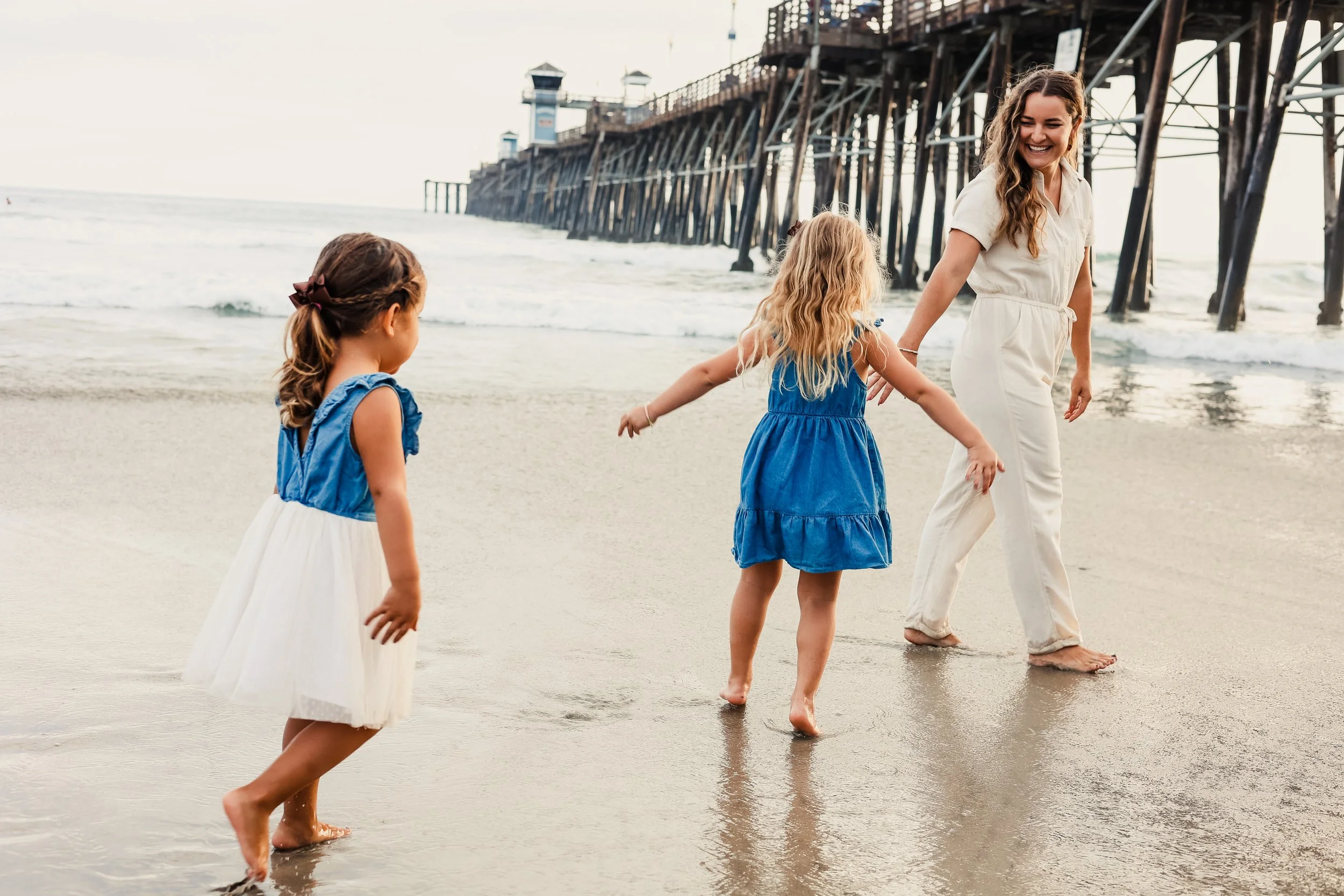 A woman and two young girls playing on the beach near a pier, with waves and the ocean in the background.