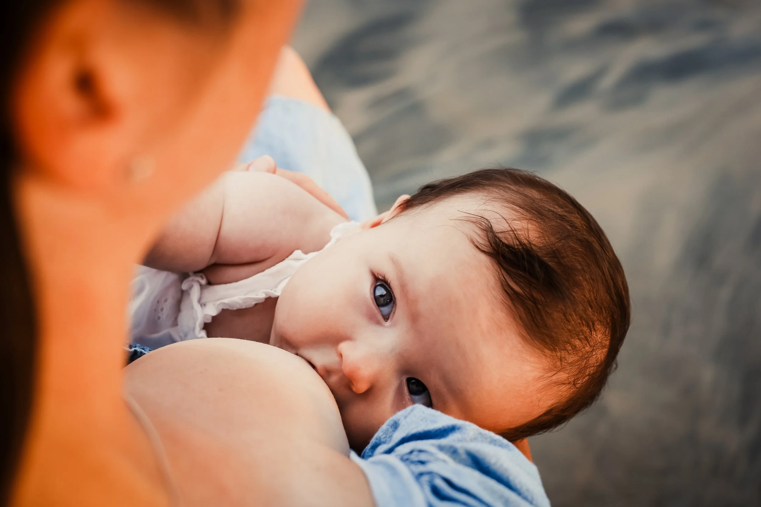A baby with brown hair and blue eyes lying on an adult's chest, breastfeeding outdoors, with a blurry background of water.