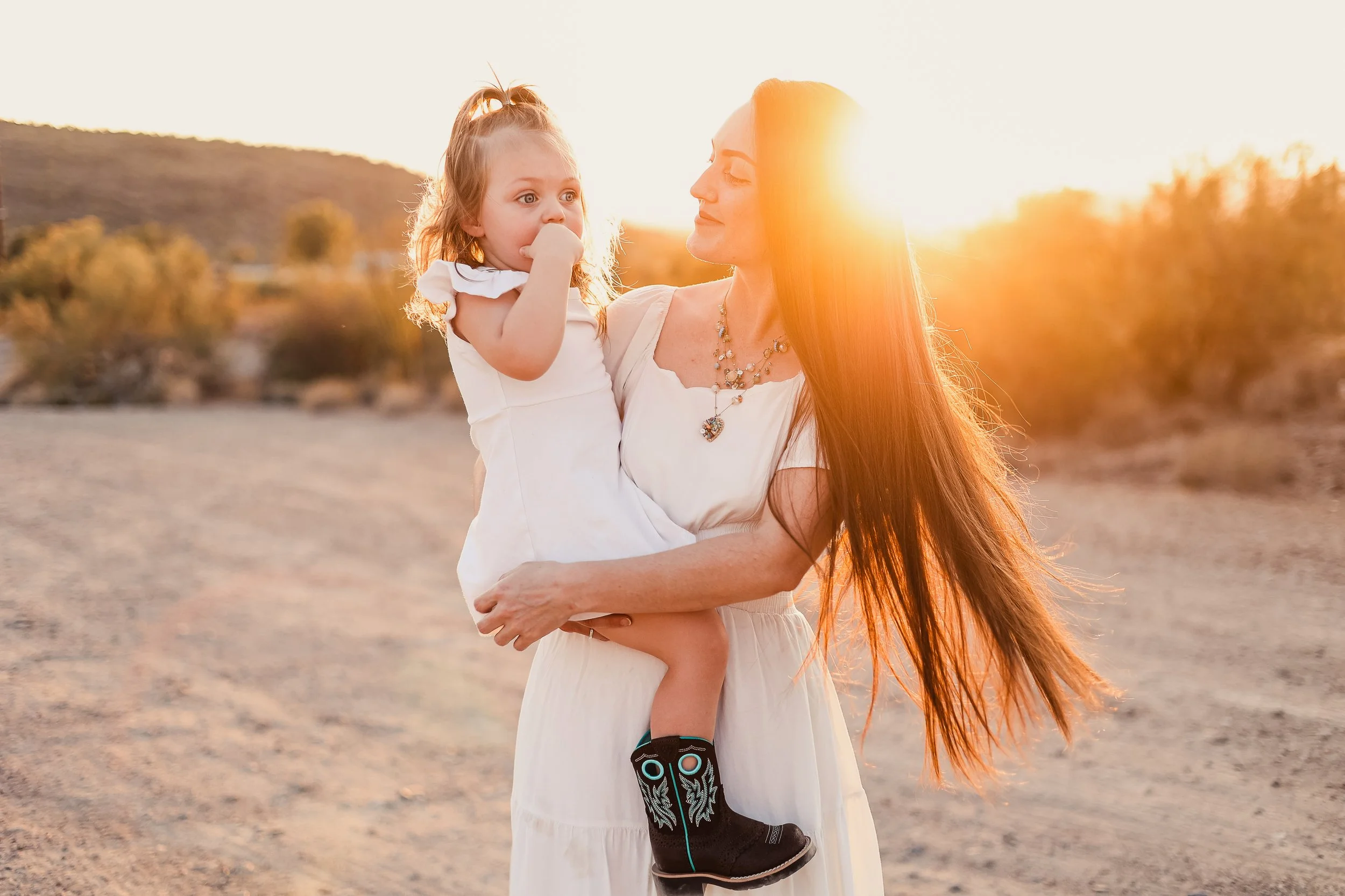 A woman holding a young girl on her arm outdoors at sunset, with mountains and bushes in the background.