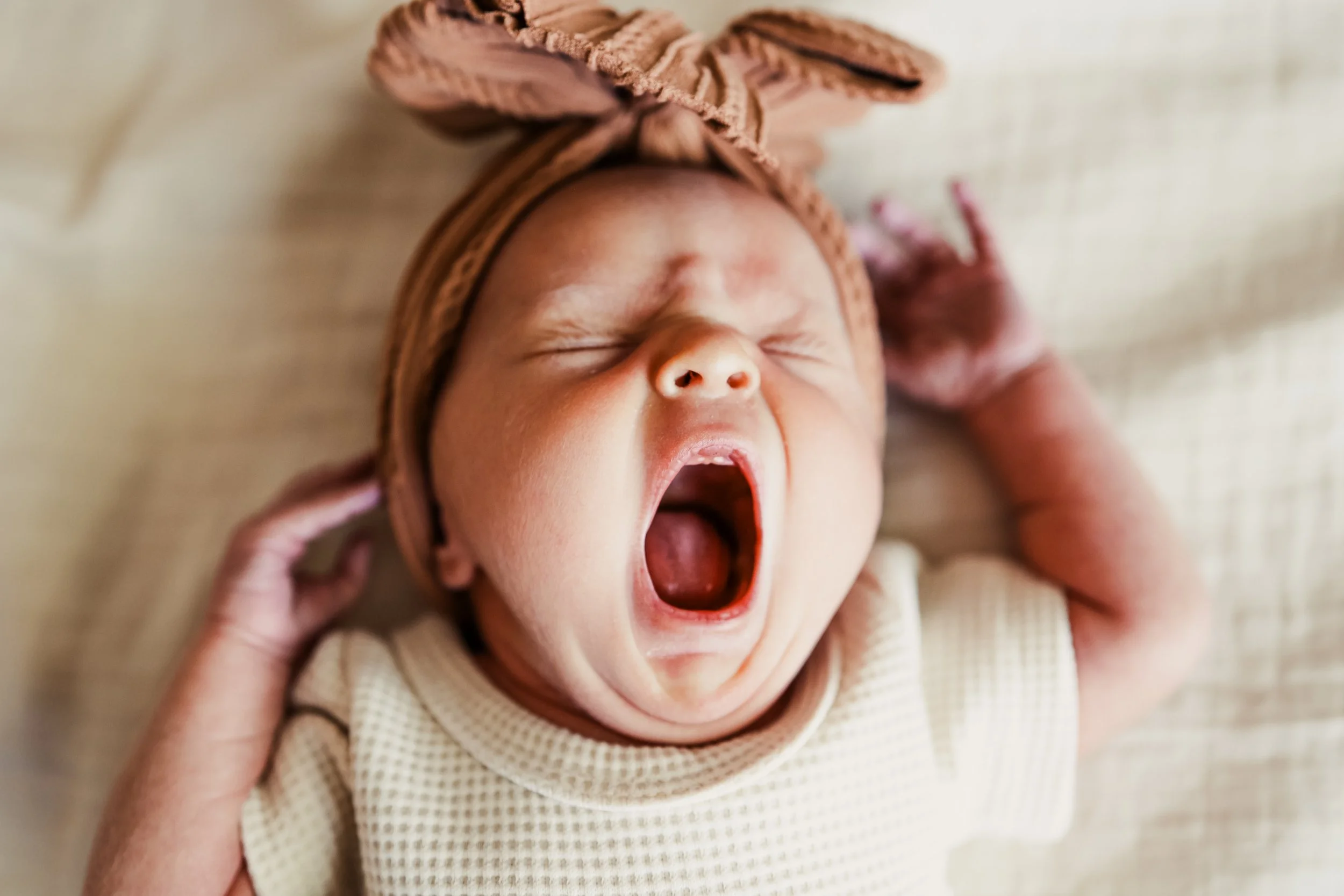 A newborn baby yawning with eyes closed, wearing a beige outfit and a brown headband with a bow, lying on a beige surface.