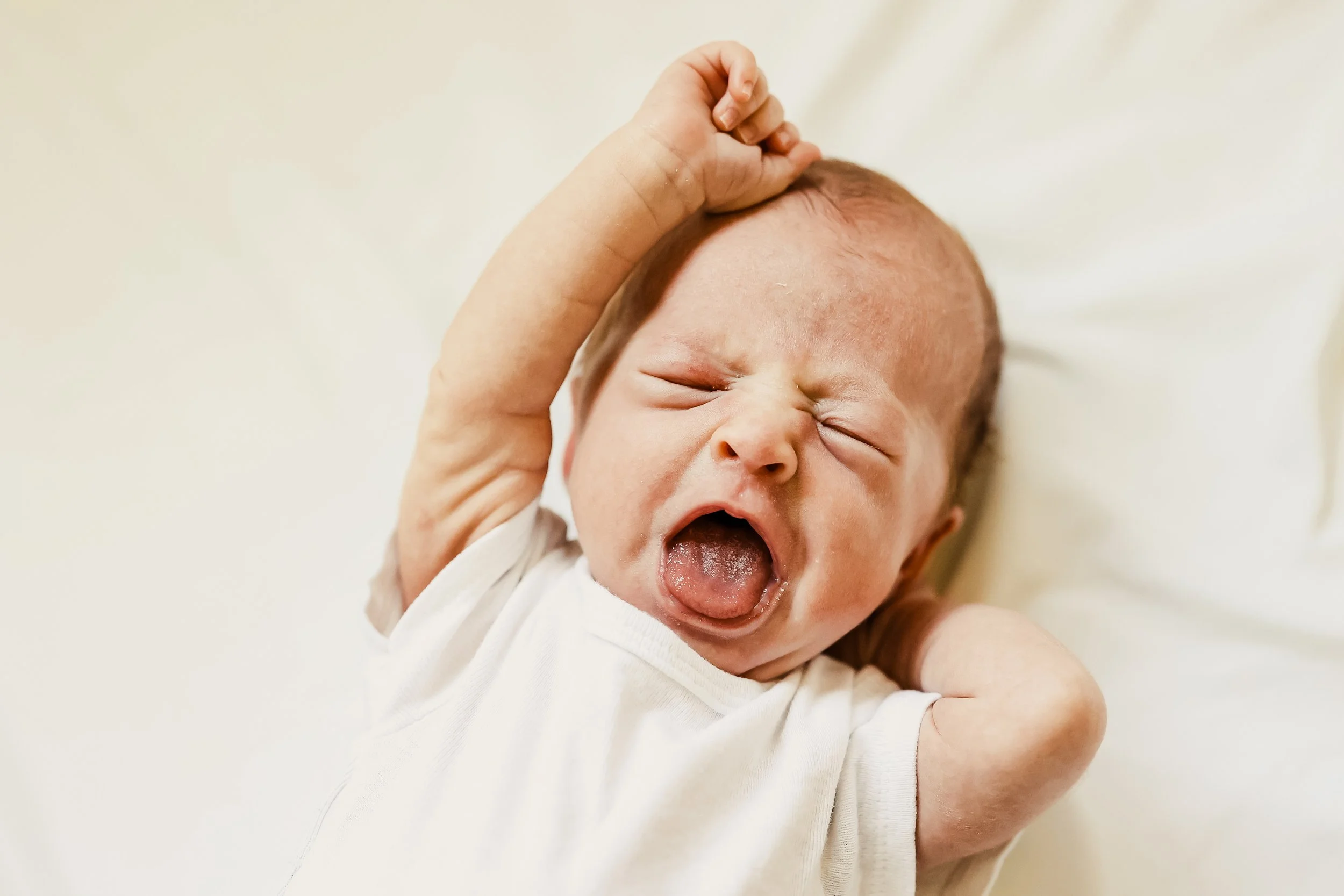 A newborn baby yawning with eyes closed, lying on a white surface, wearing a white onesie, and raising one arm.
