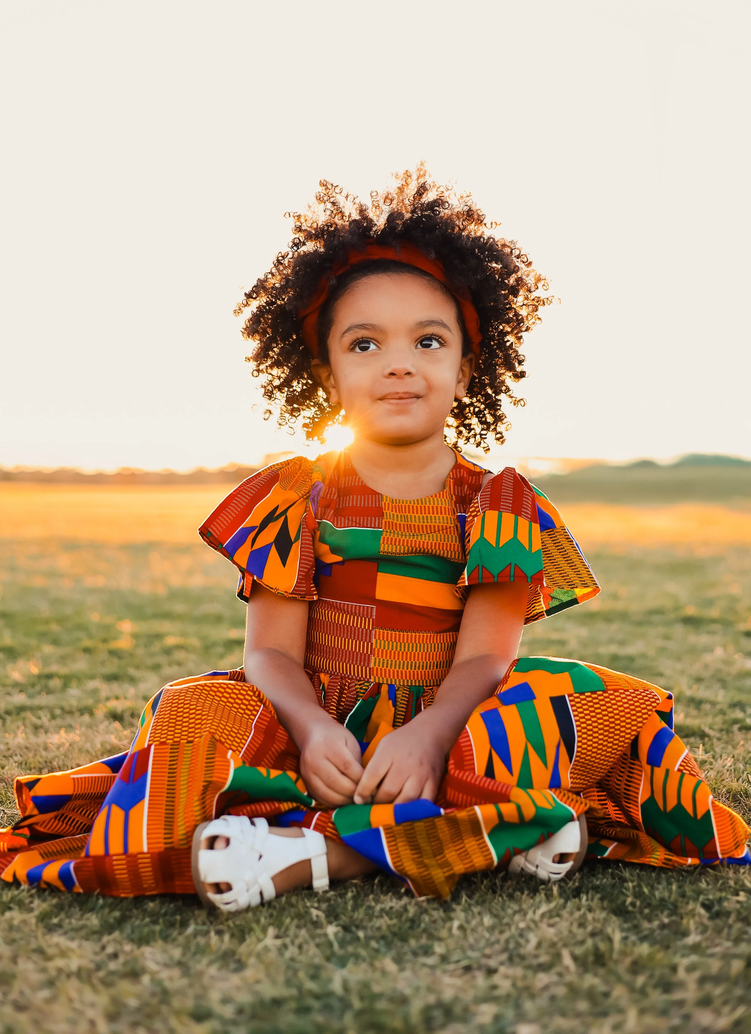 A young girl with curly hair sitting on grass at sunset, wearing a colorful patterned dress and white sandals.